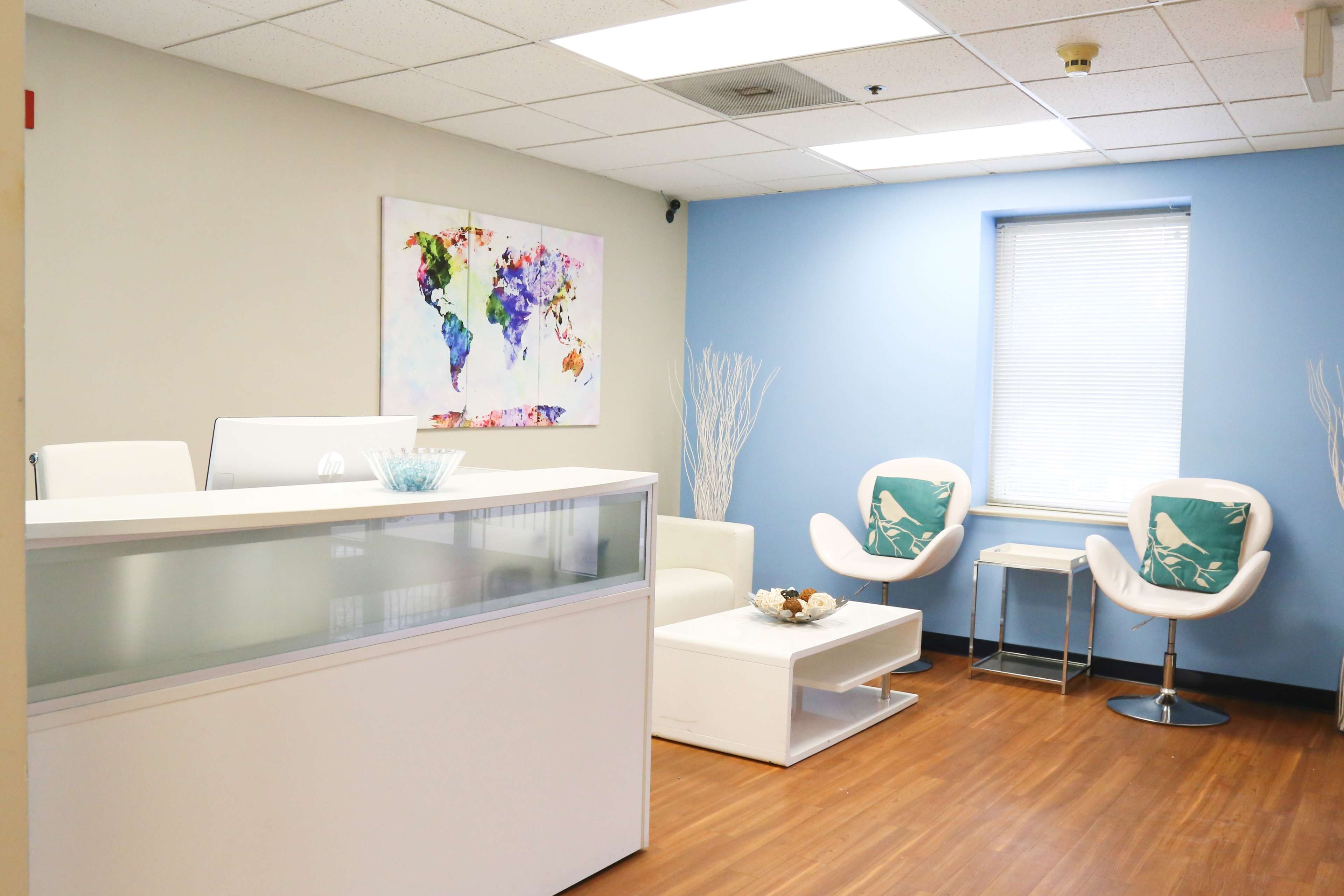A modern office reception area with a white desk, two chairs, a coffee table, and a colorful world map painting on the wall.