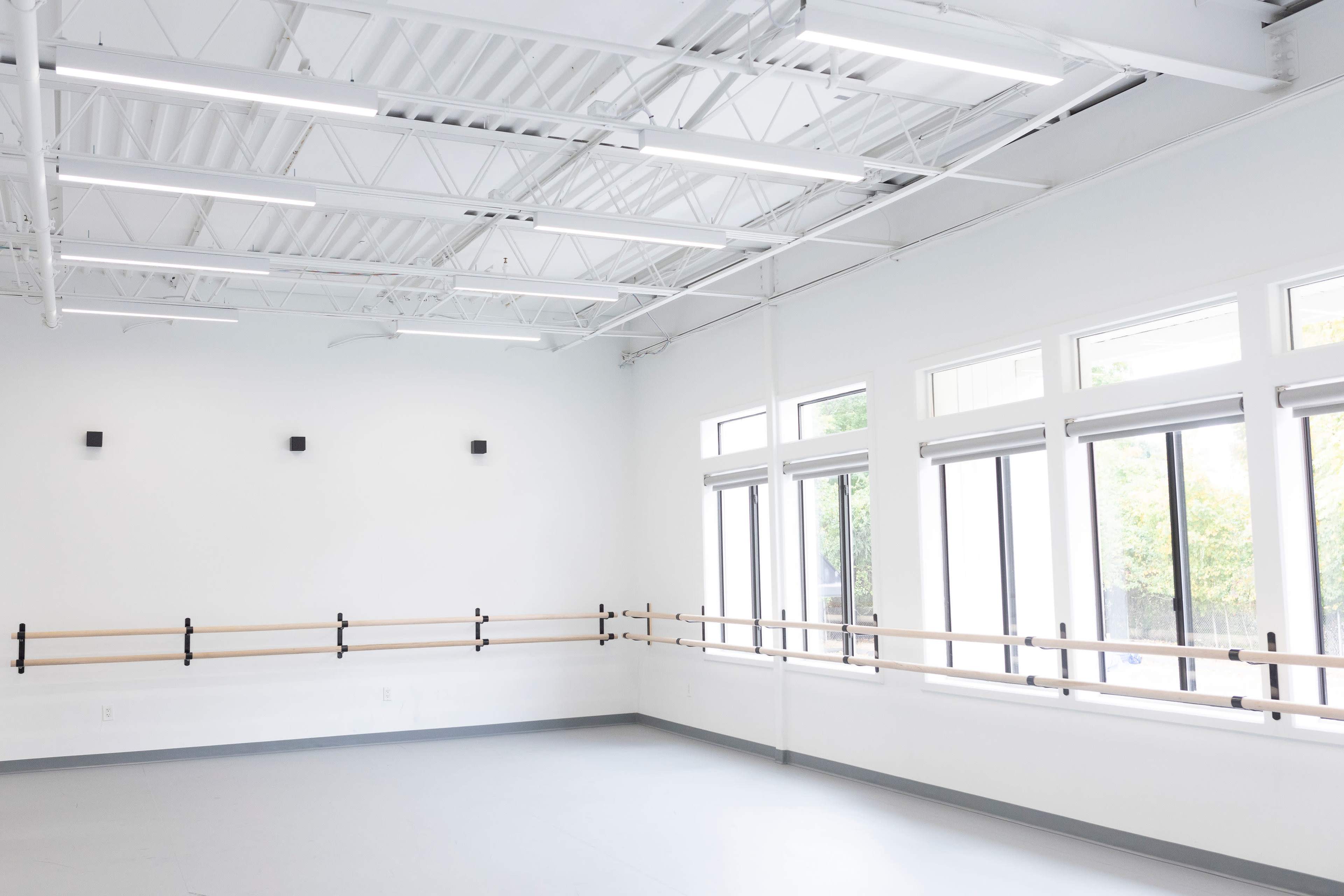 The image shows a spacious, well-lit dance studio with large windows and a wall-mounted ballet barre.
