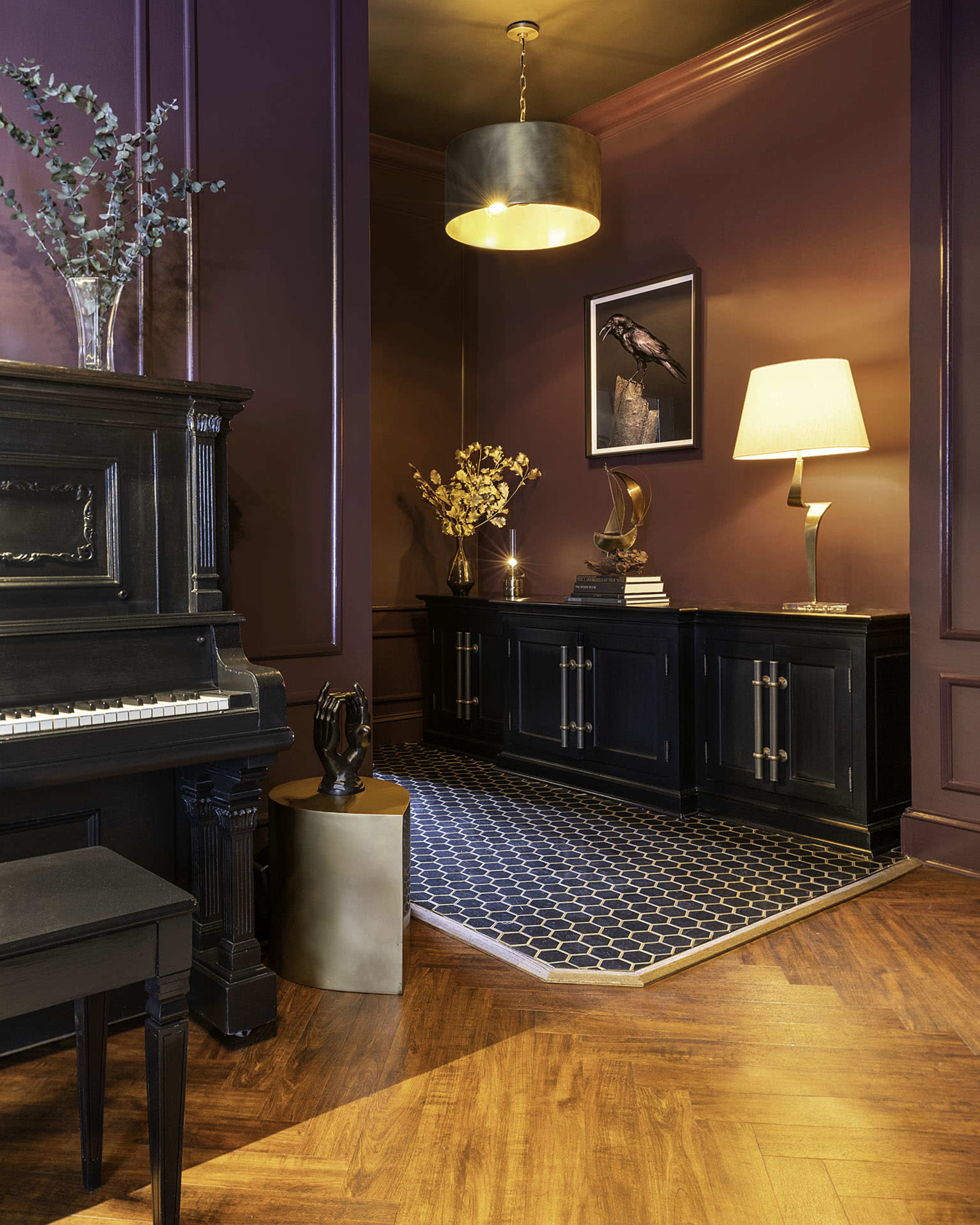 A dark-hued corner of a room features a black piano, a sideboard with decorative objects, and a geometric area rug on a wooden floor.
