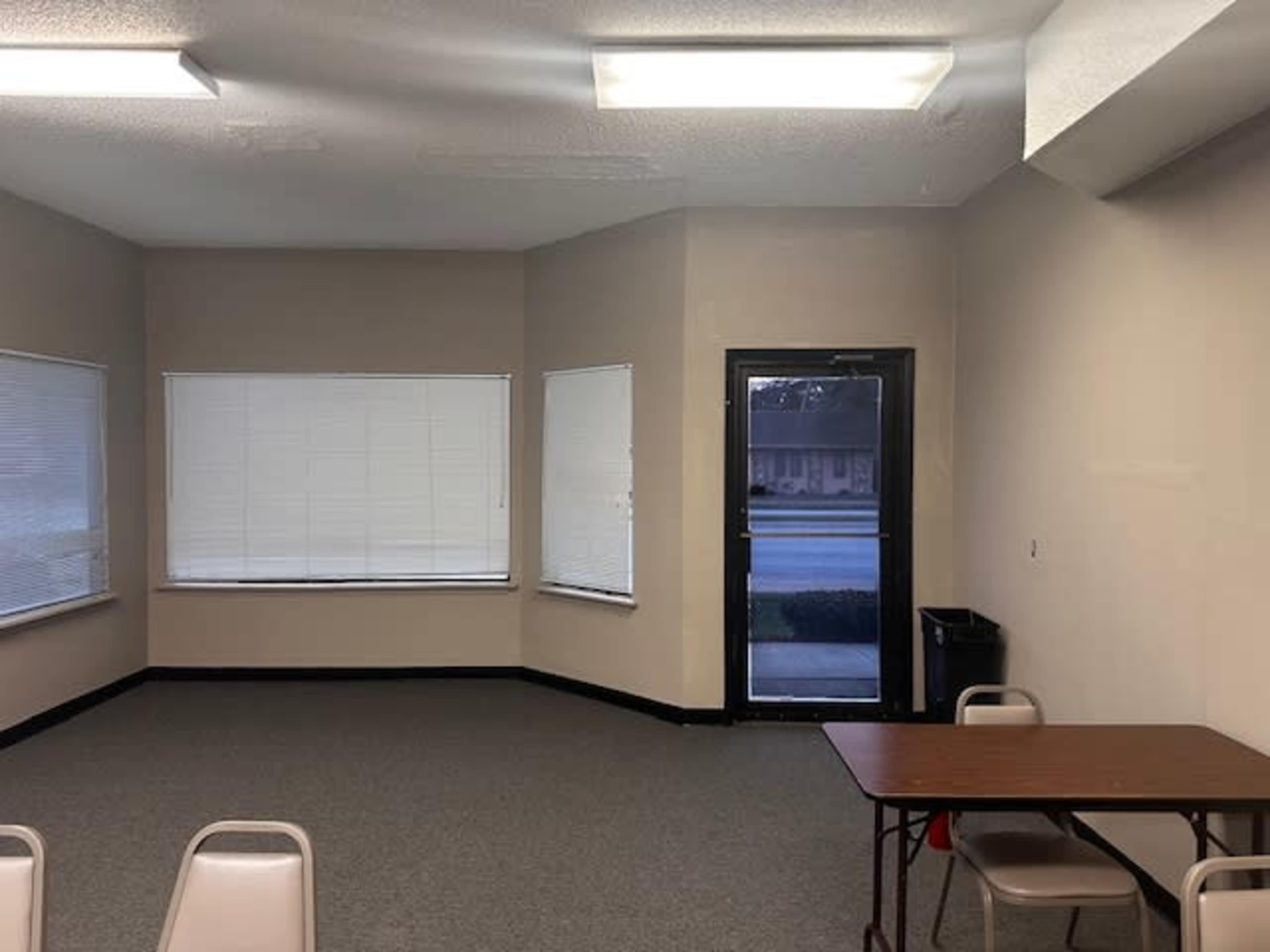 The image shows a sparsely furnished room with a table and chairs, large windows, and a door leading outside.