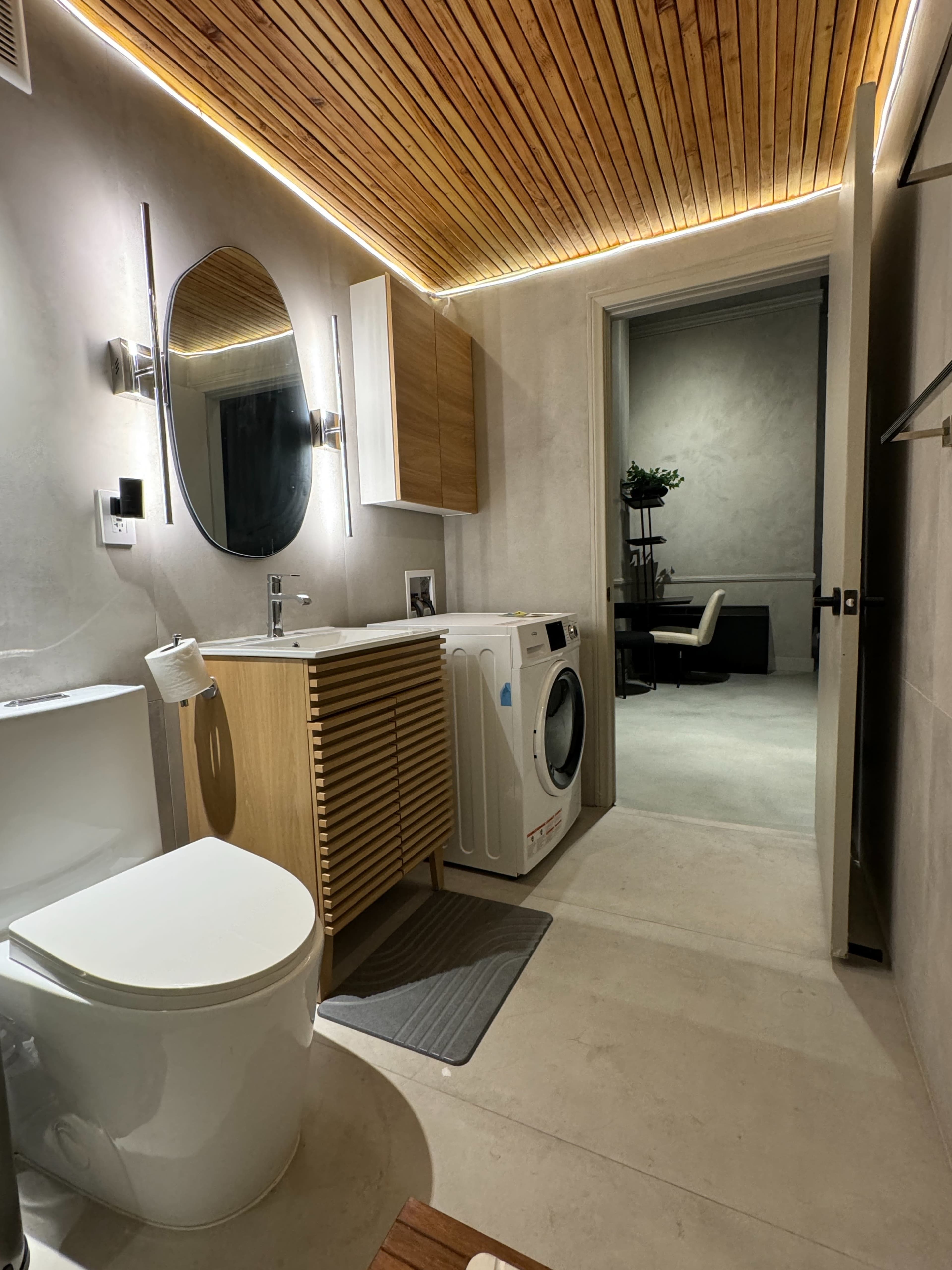 The image shows a modern bathroom featuring a toilet, a sink with a wooden vanity, a washing machine, and a doorway leading to another room.