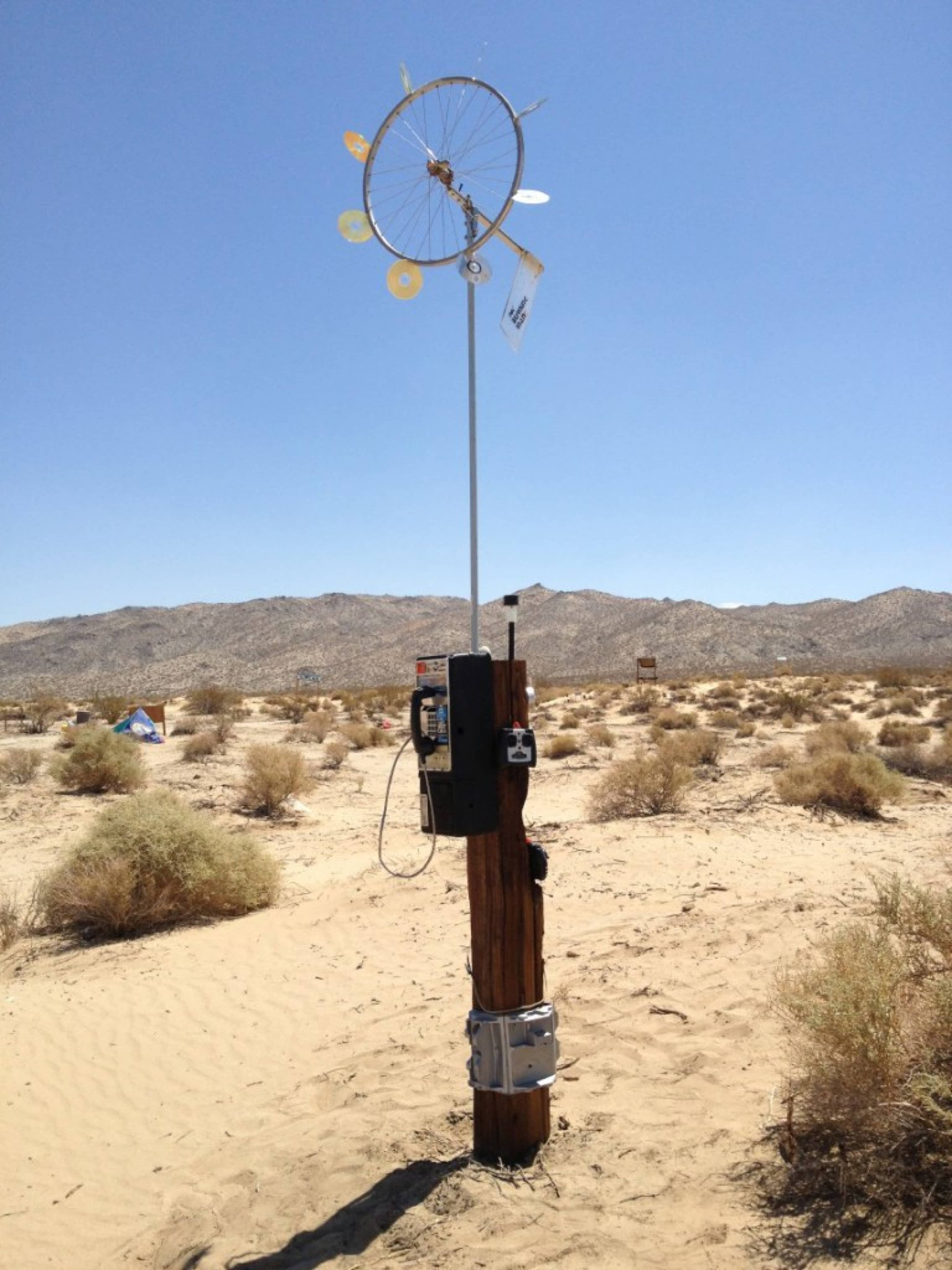 A wooden post in a desert landscape displays a wind-catching device made of discs and a telephone booth attached to its side.