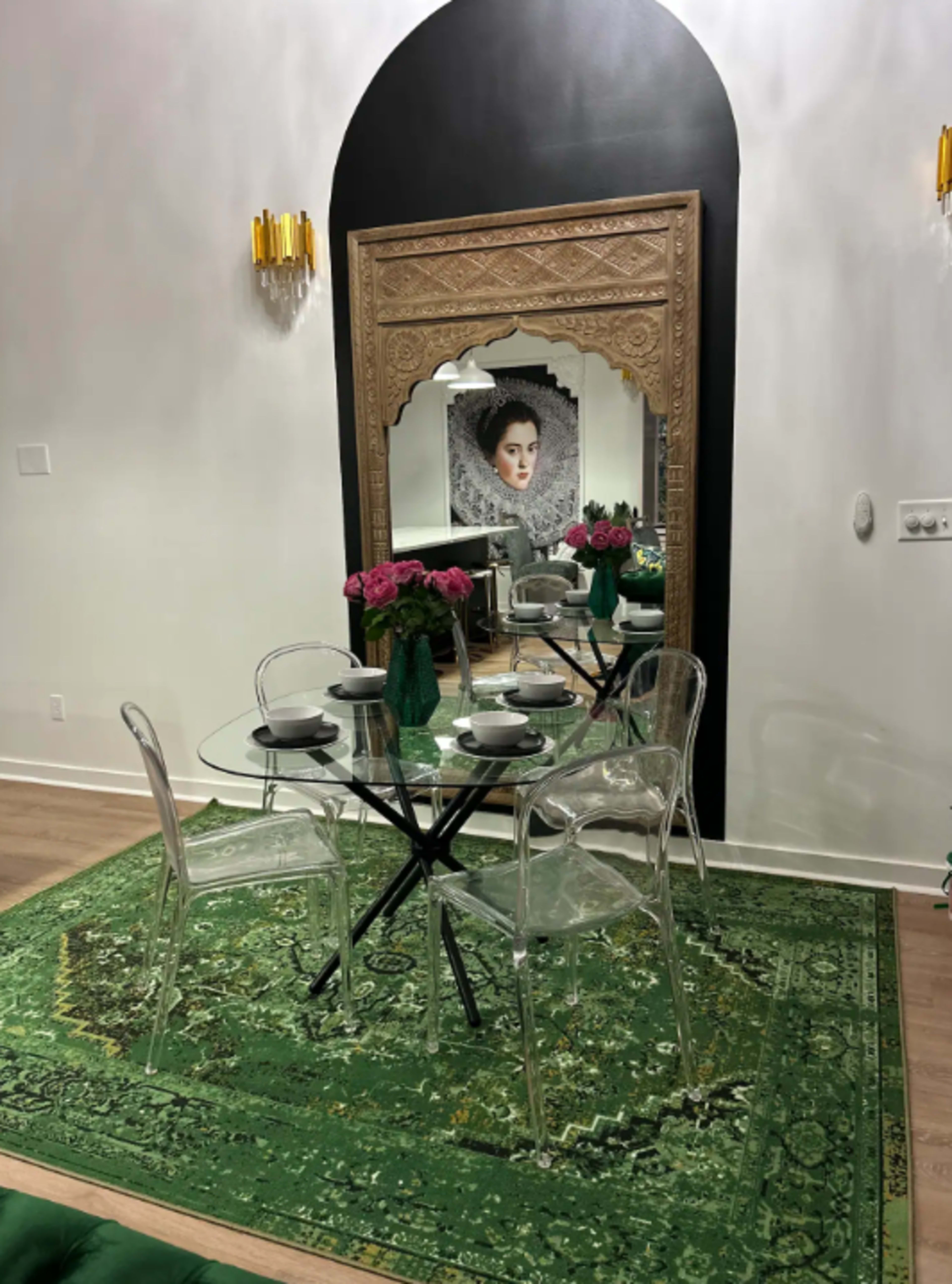A small dining area features a glass table with transparent chairs, surrounded by a green patterned rug and a large decorative mirror reflecting a framed portrait.