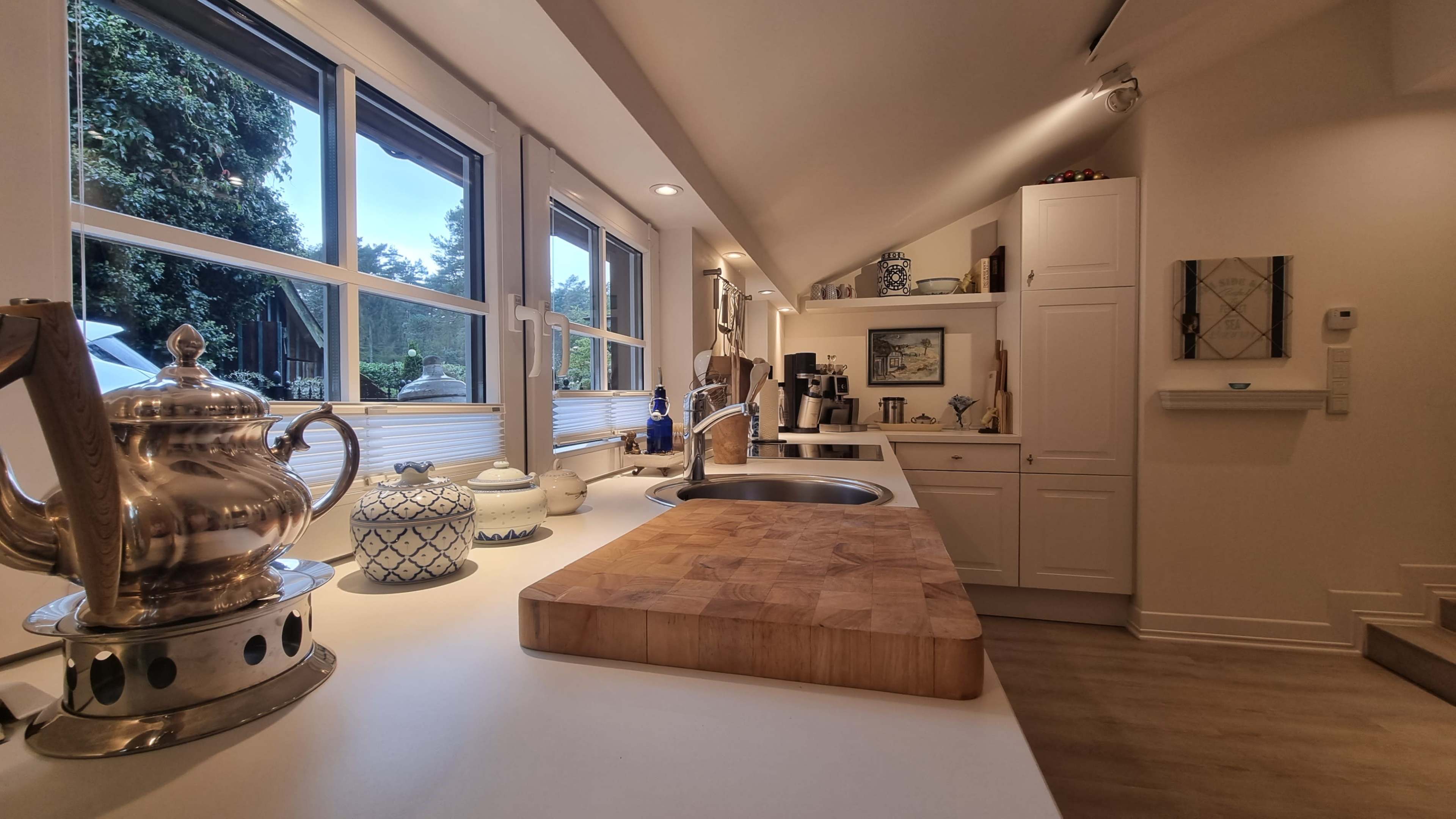The image shows a modern kitchen with a wooden cutting board on a white countertop, silver teapots, and large windows letting in natural light.