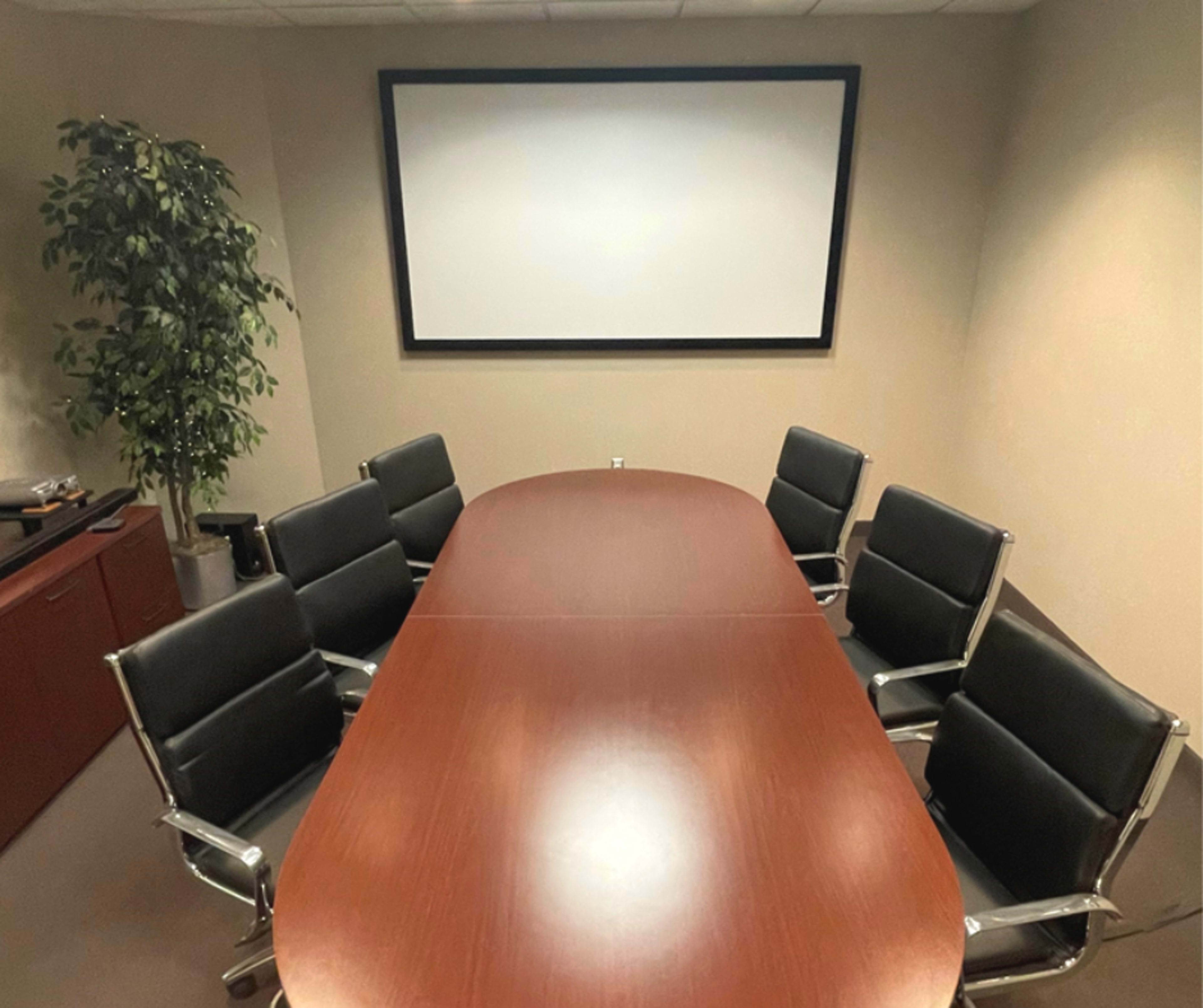 A wooden conference table is placed in a meeting room surrounded by six black swivel chairs, with a large screen mounted on the wall.