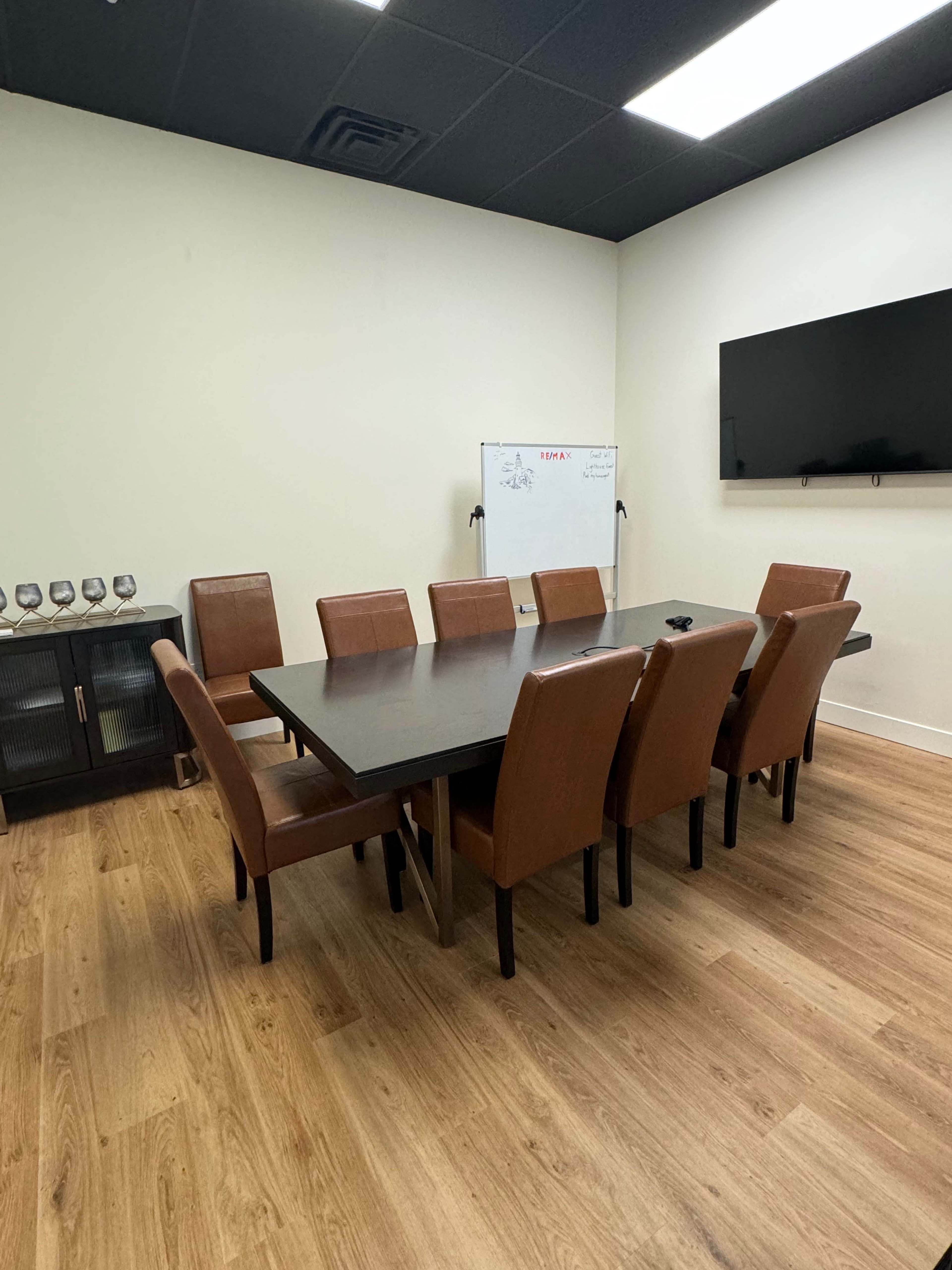 A conference room with a large table surrounded by eight brown chairs, a whiteboard, and a wall-mounted television.