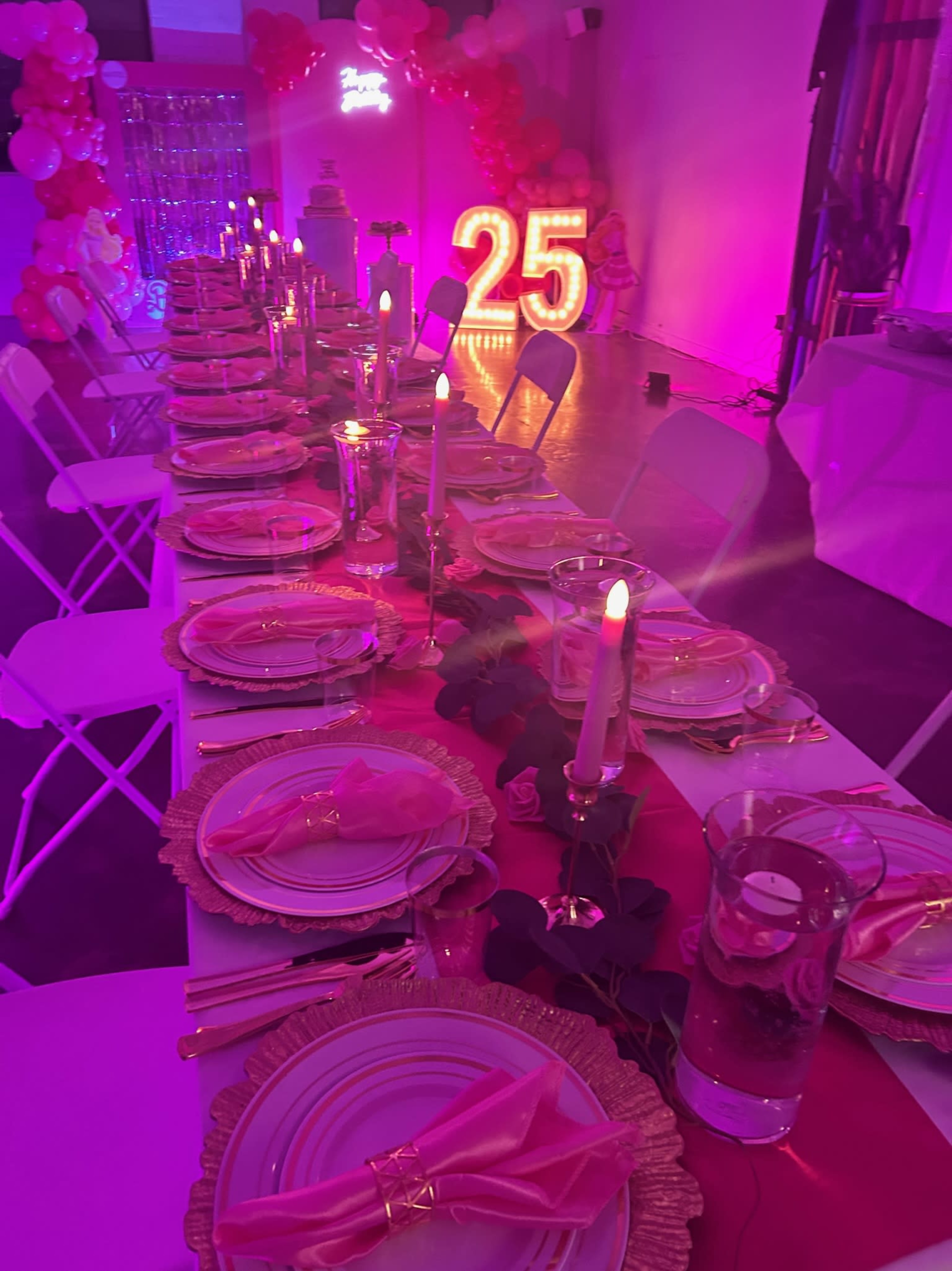 A long dining table is set with pink plates and candles, decorated with a pink runner, and illuminated by large light-up numbers "25" against a backdrop of balloons.