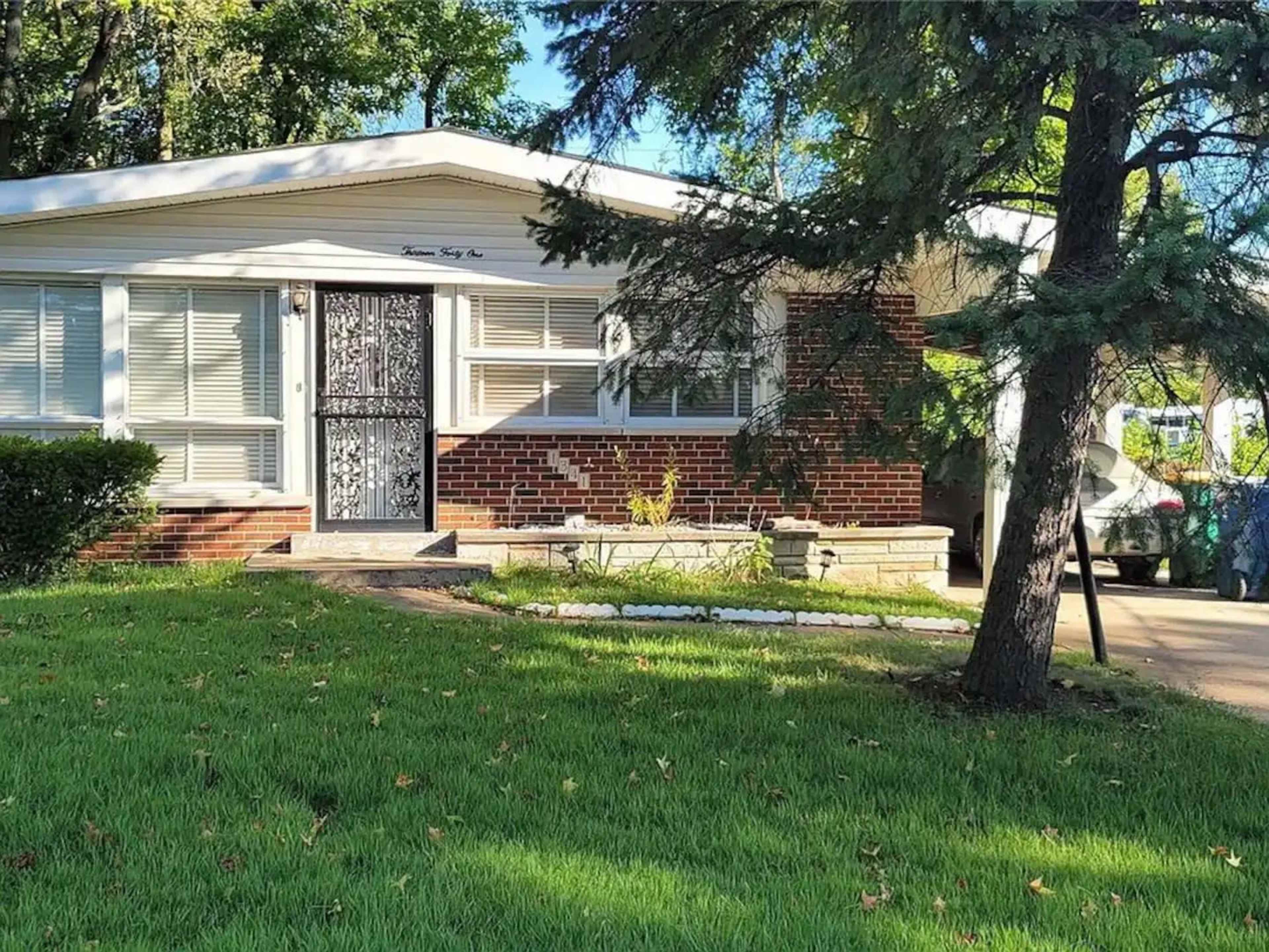 A single-story brick house with white window shutters and a black wrought-iron front door, surrounded by green grass and trees.