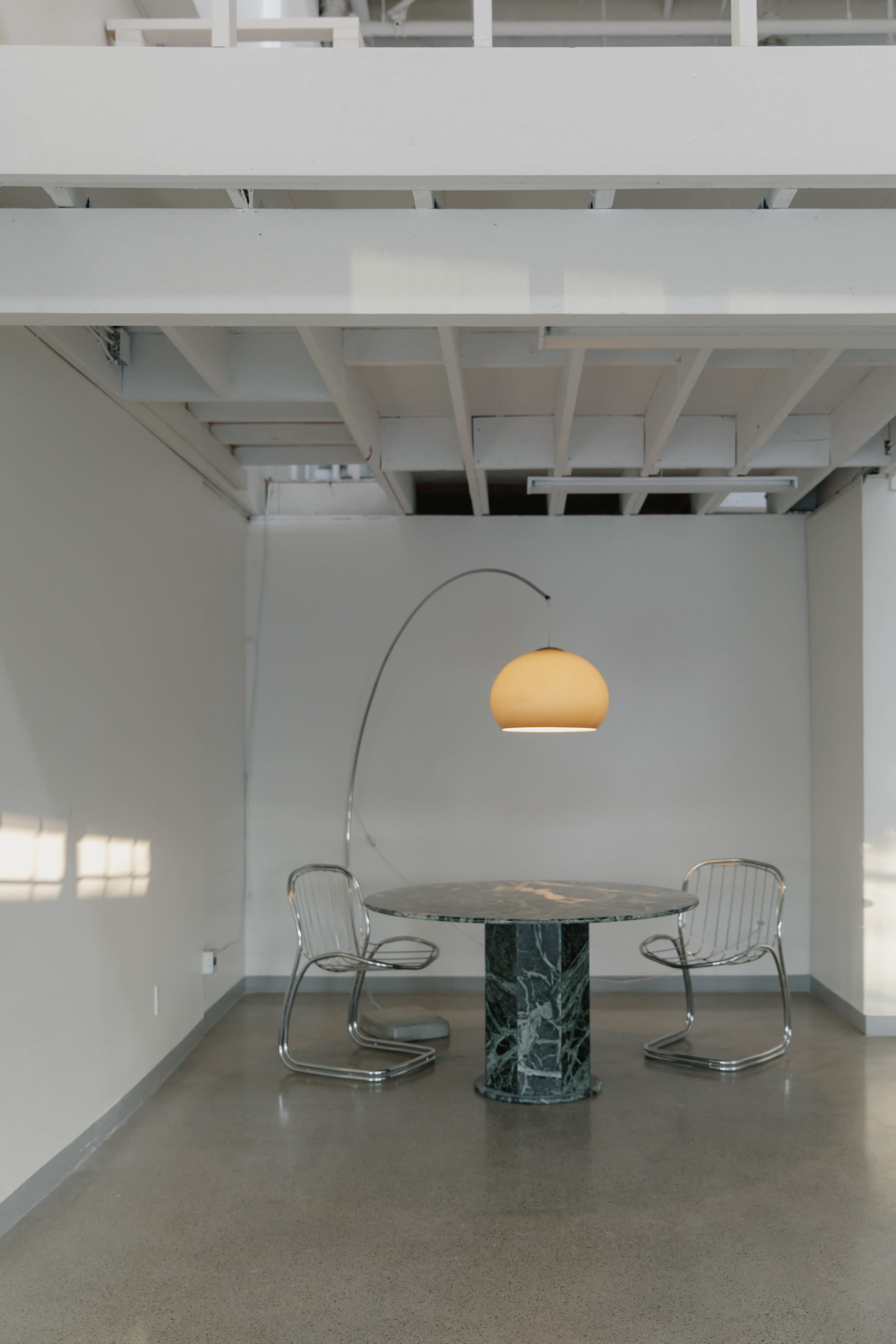 A round table with a marble base and two metal chairs is illuminated by a large pendant light in a minimalist interior space.