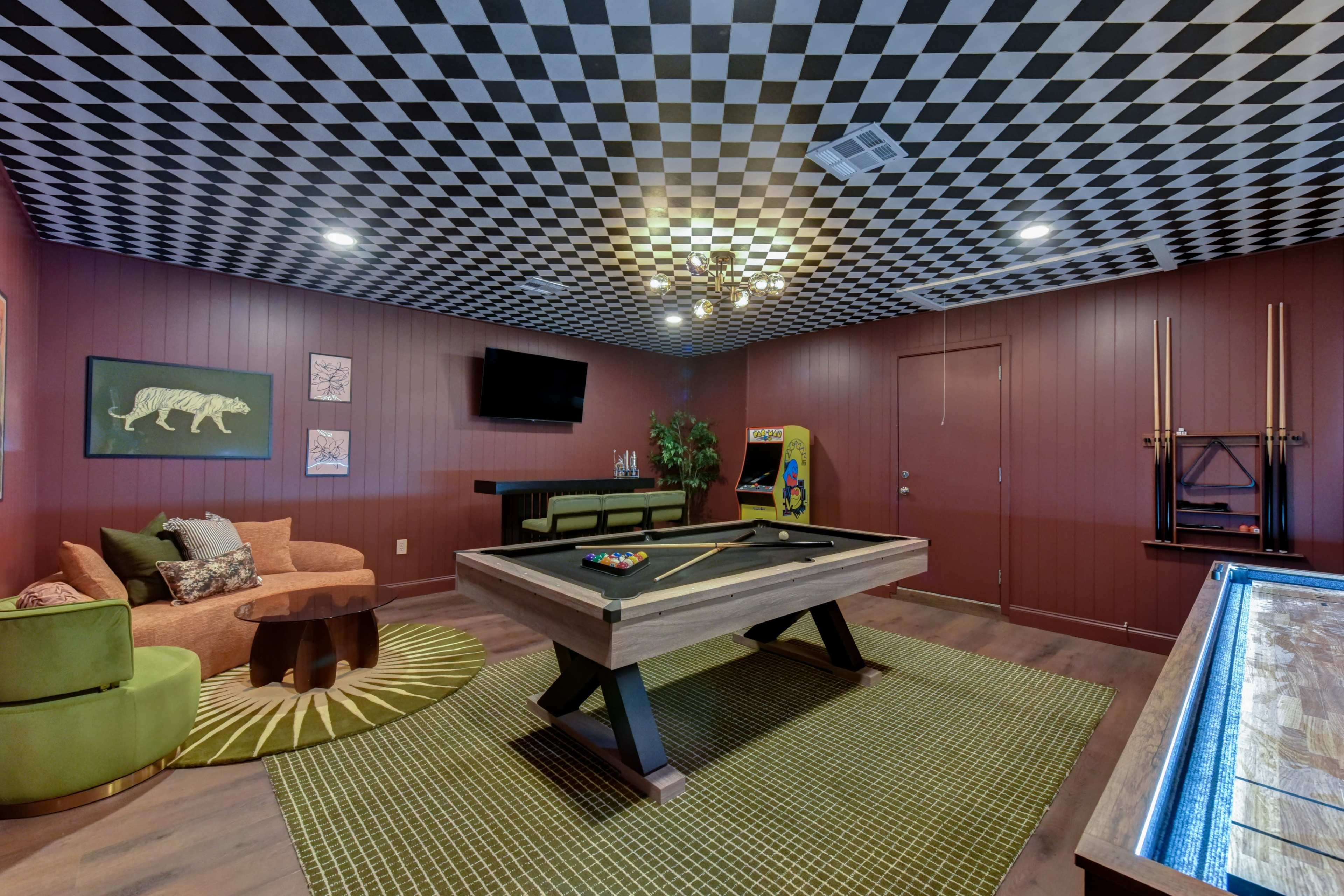 The image shows a stylish game room featuring a pool table, a green area rug, and a checkerboard ceiling, with a seating area and a small bar in the corner.