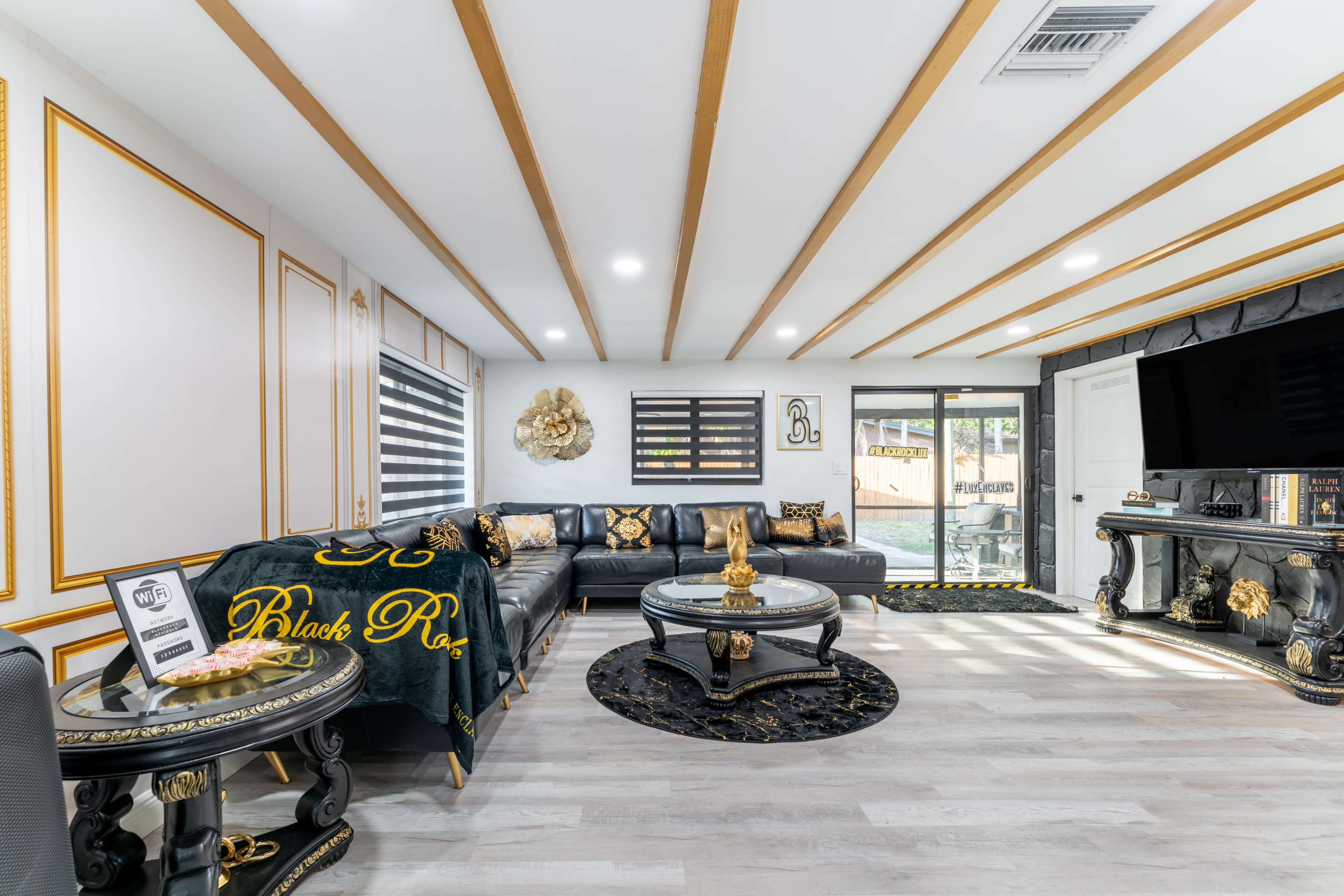 The living room features a modern design with a black leather sectional, a round glass coffee table, and decorative gold accents on the walls and furniture.