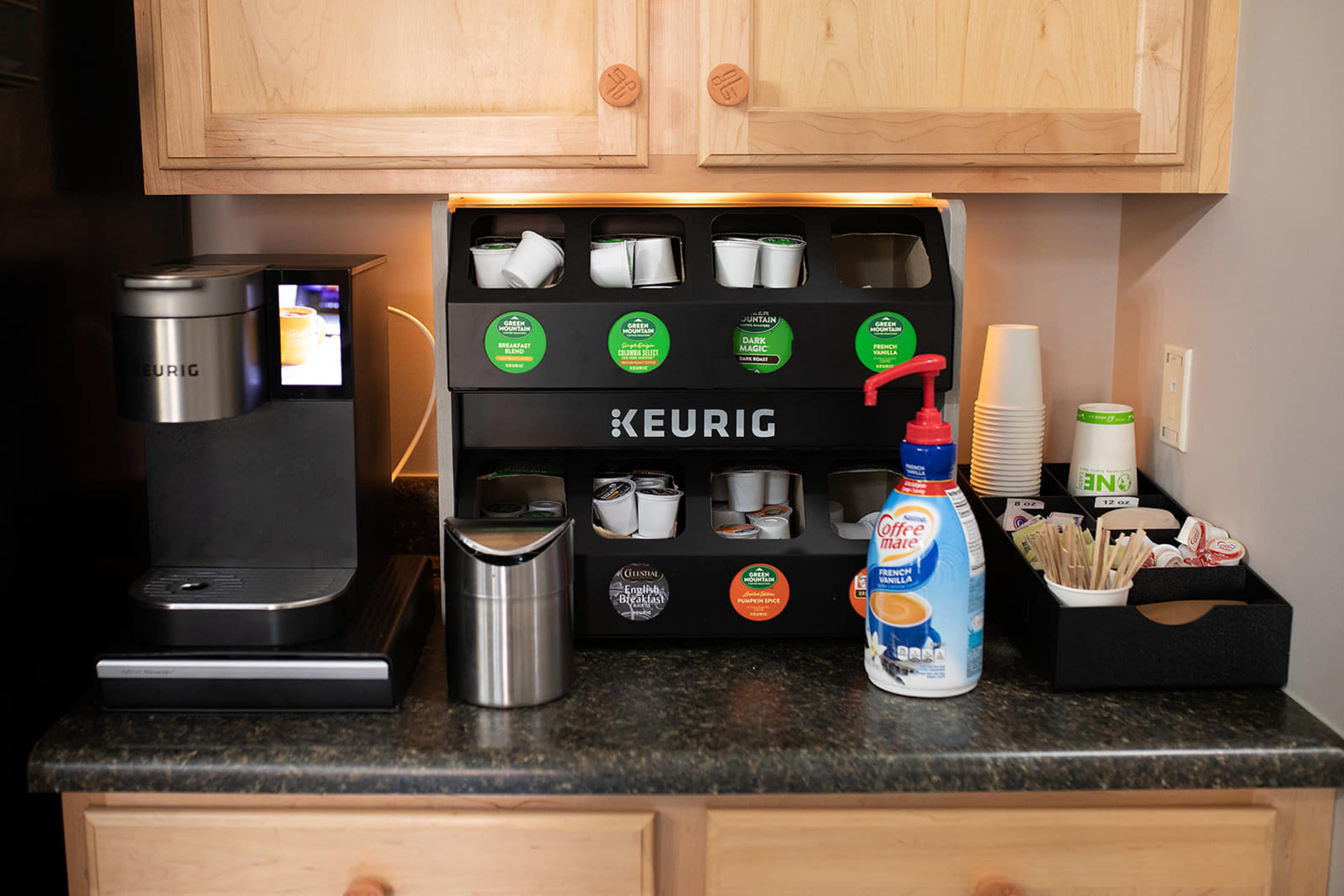 The image shows a Keurig coffee machine alongside a storage unit for coffee pods, cups, and creamers on a countertop.