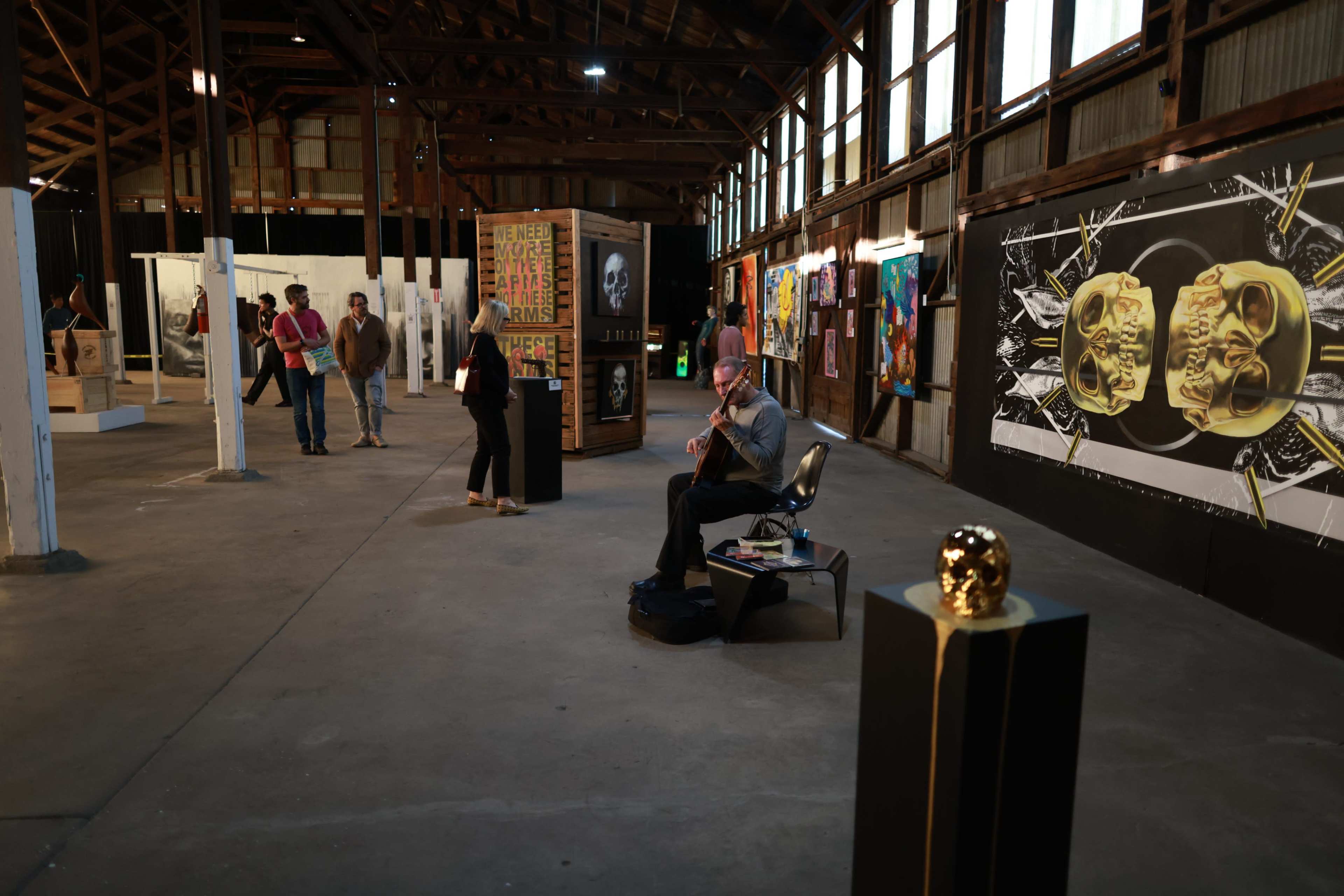 An art gallery features various artworks along the walls, with visitors exploring the space while a musician plays guitar in the center.
