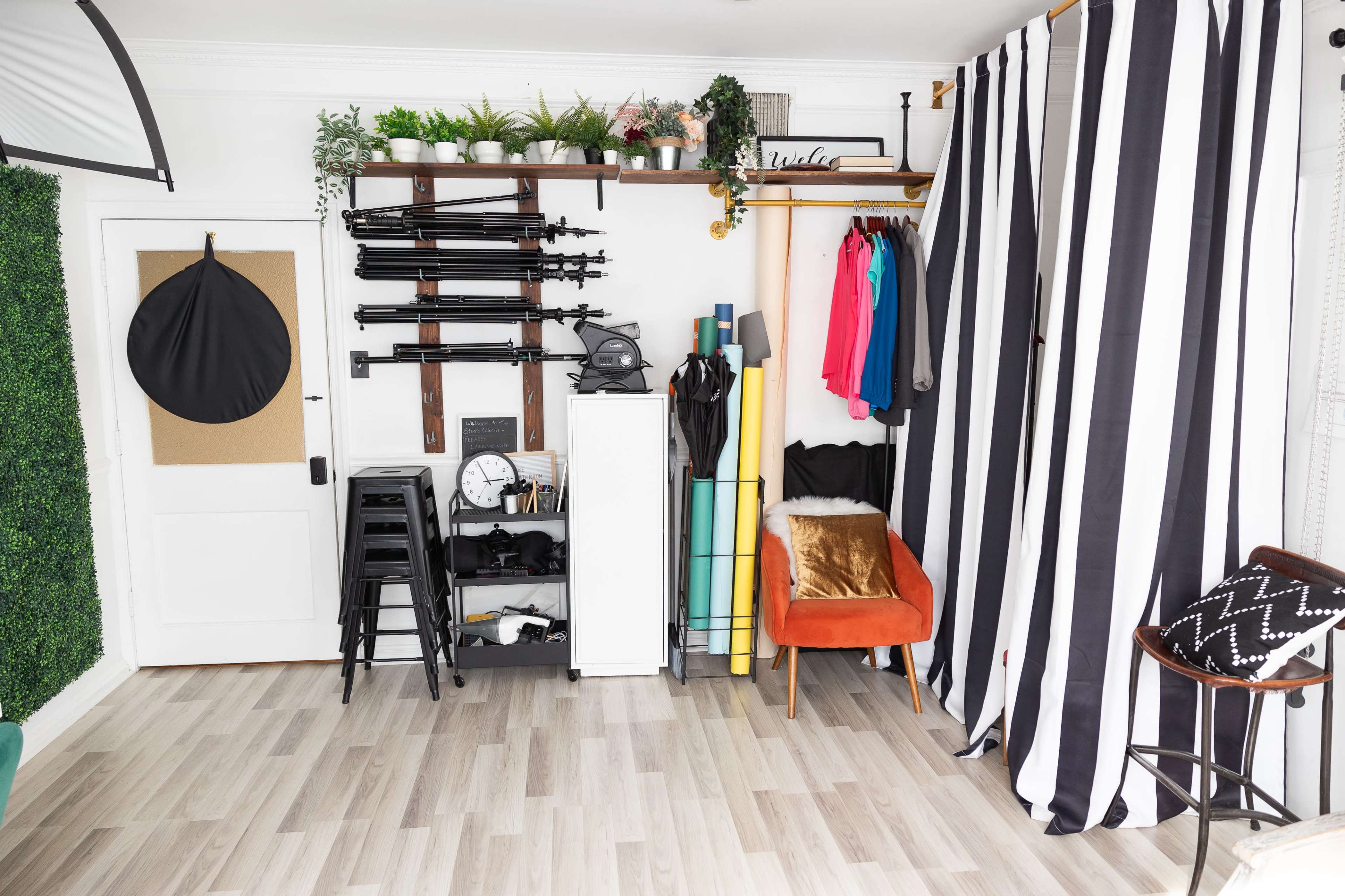 A studio space equipped with various props, clothing racks, and storage items against a backdrop of striped curtains and greenery.