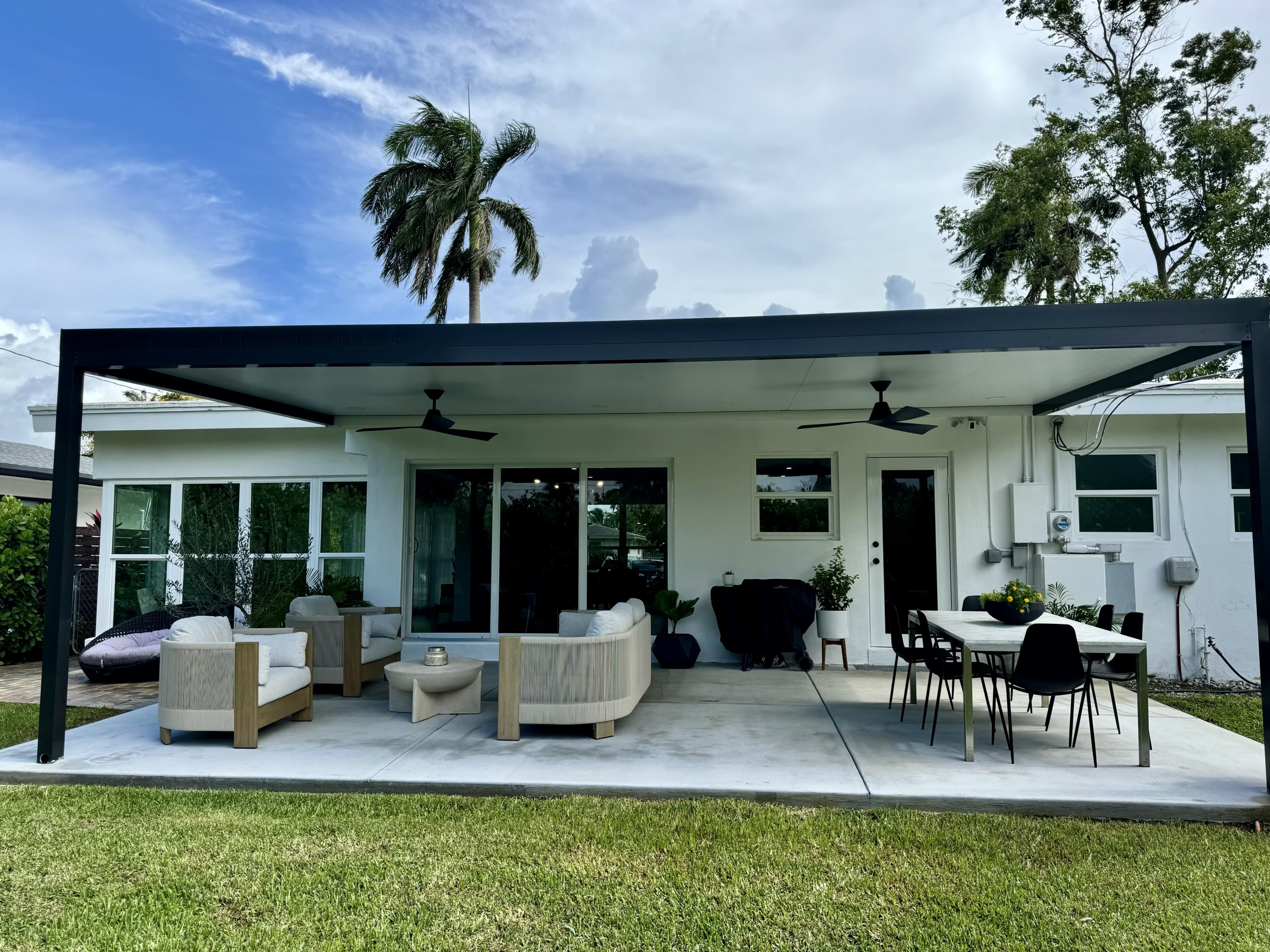 The image shows a covered outdoor patio area with a seating arrangement and dining table, surrounded by grass and palm trees.