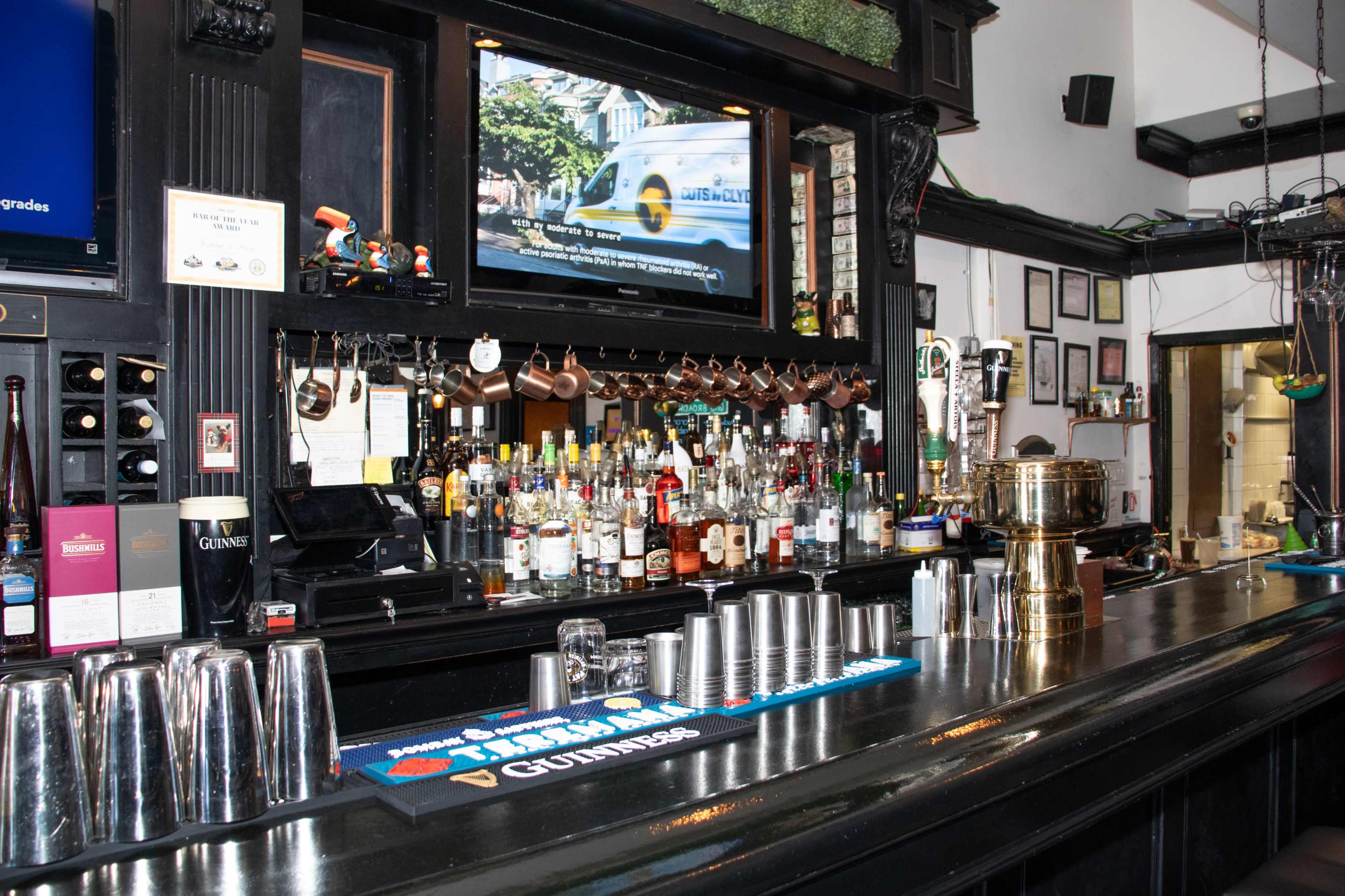 A bar counter is lined with various bottles of alcohol, a television displays a program above, and several metal shakers are neatly arranged in front.