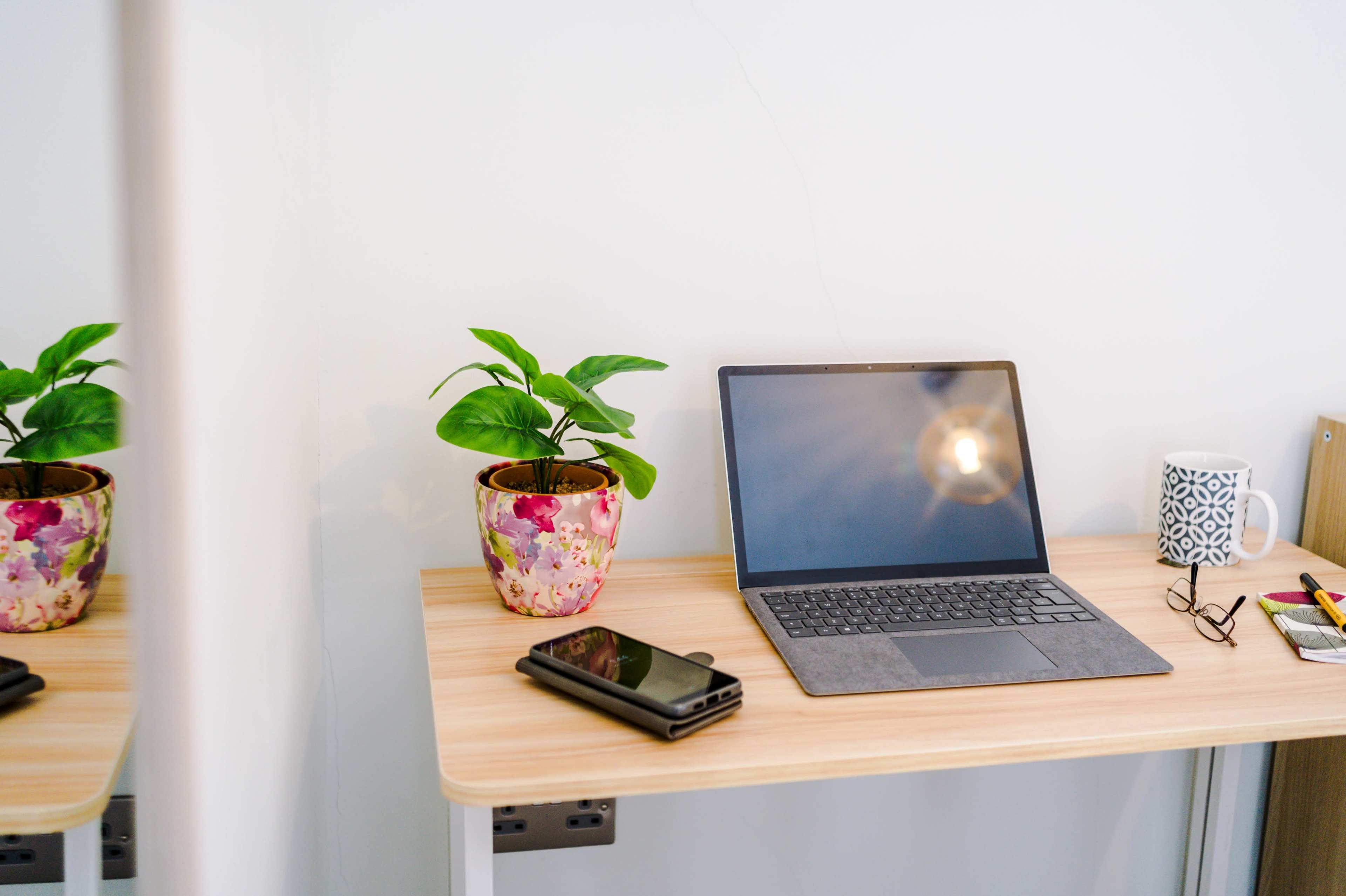 A wooden desk holds a laptop, a smartphone, a coffee mug, reading glasses, and a potted plant against a minimalist wall.