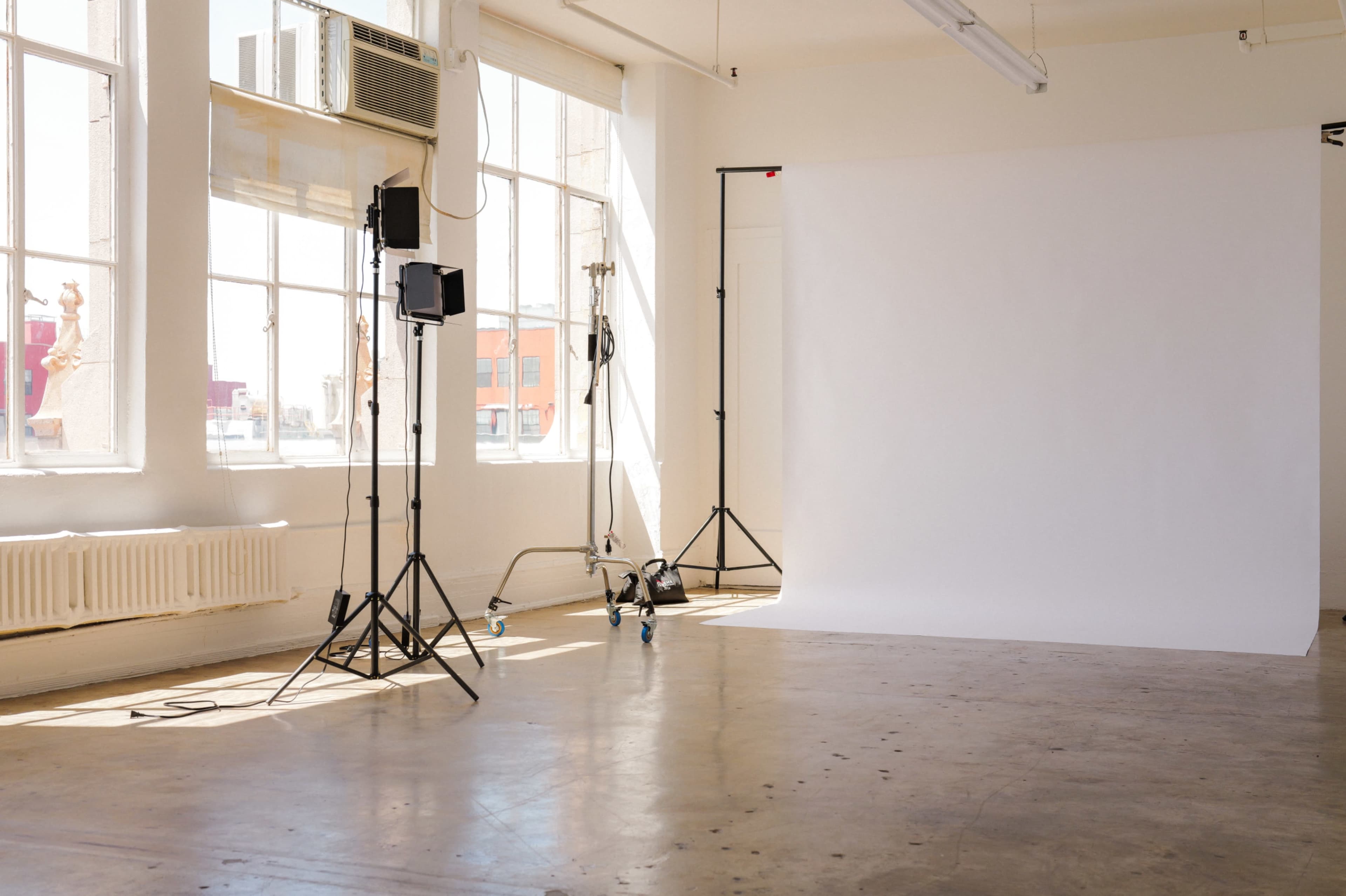 The image shows a bright, empty photography studio with large windows, a seamless backdrop, and lighting equipment set up on stands.