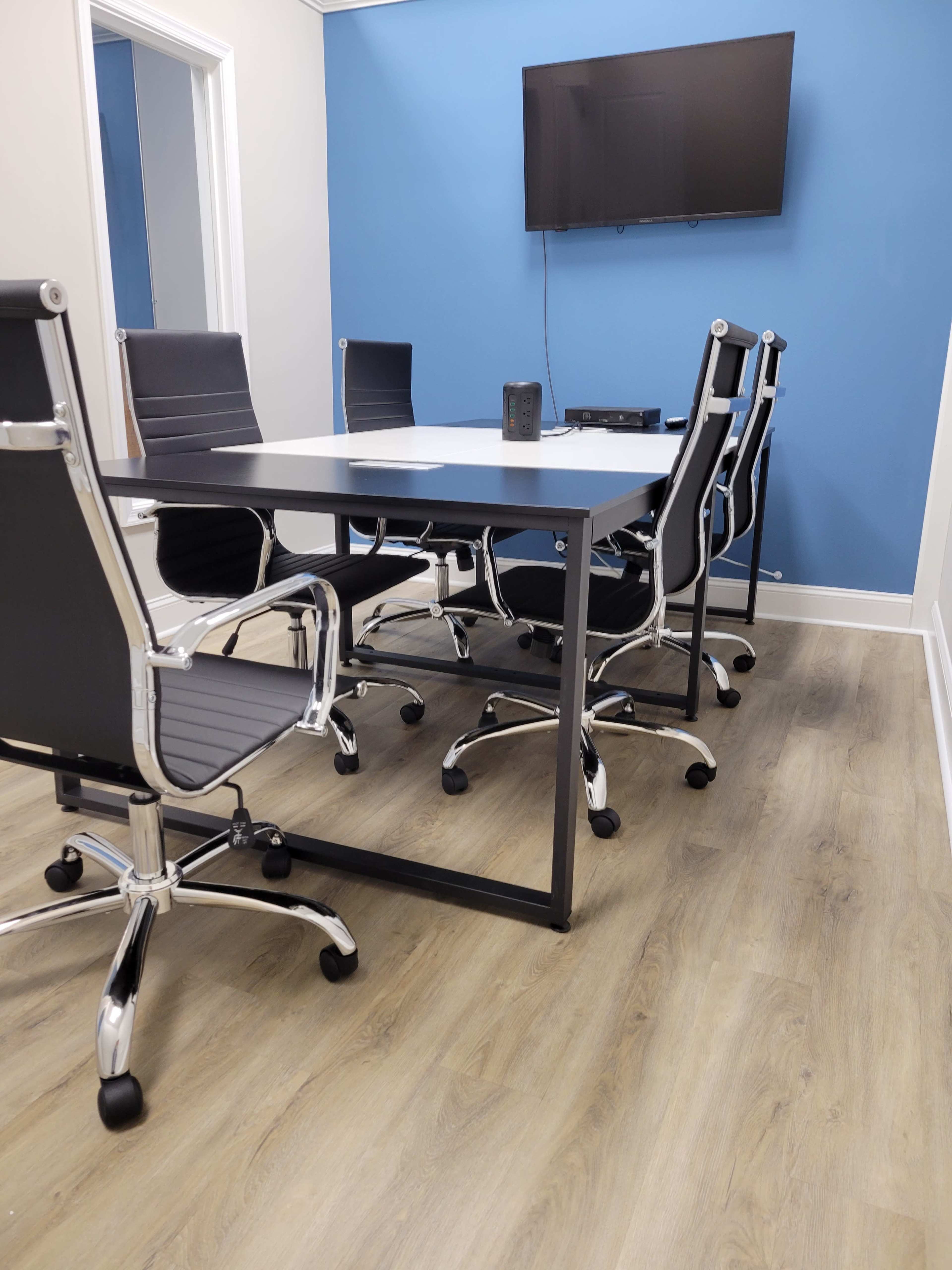A conference room features a rectangular table surrounded by chrome and black chairs, with a television mounted on a blue wall.