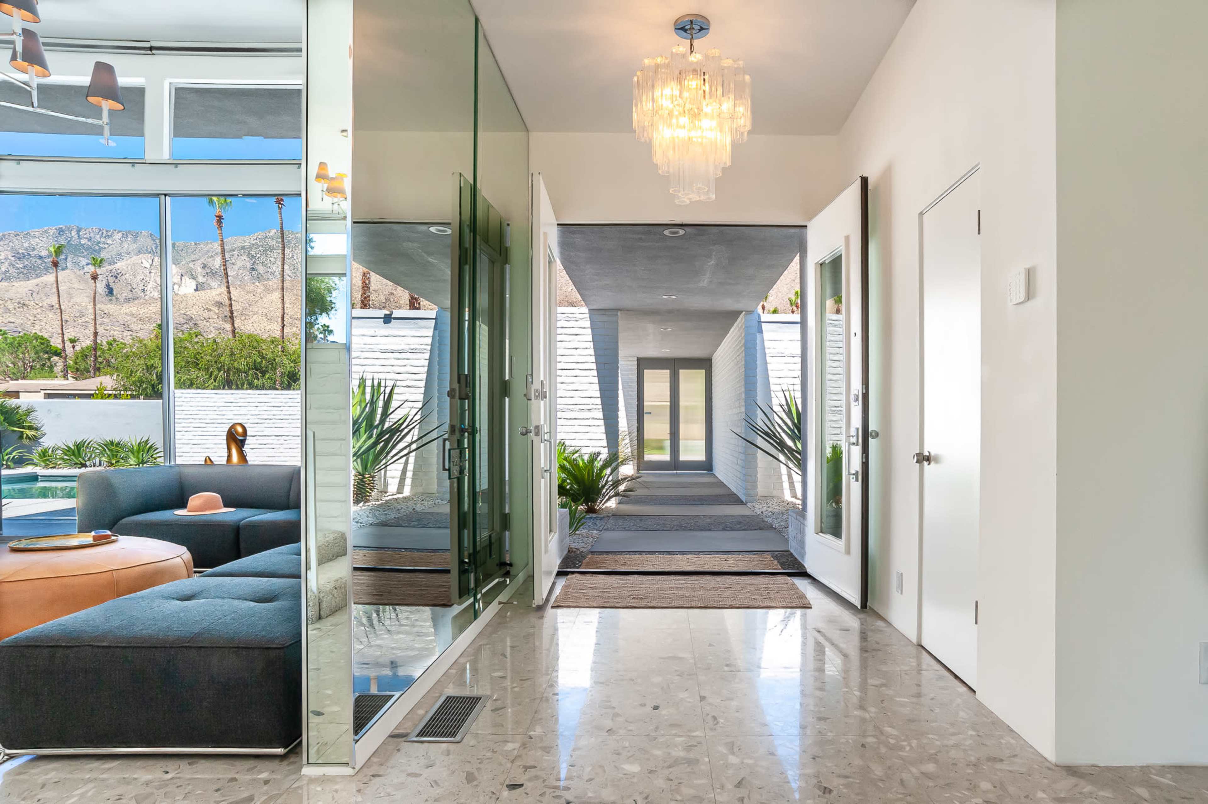 A modern entryway with large glass doors, a chandelier, and a clear view of a landscaped outdoor area framed by palm trees.