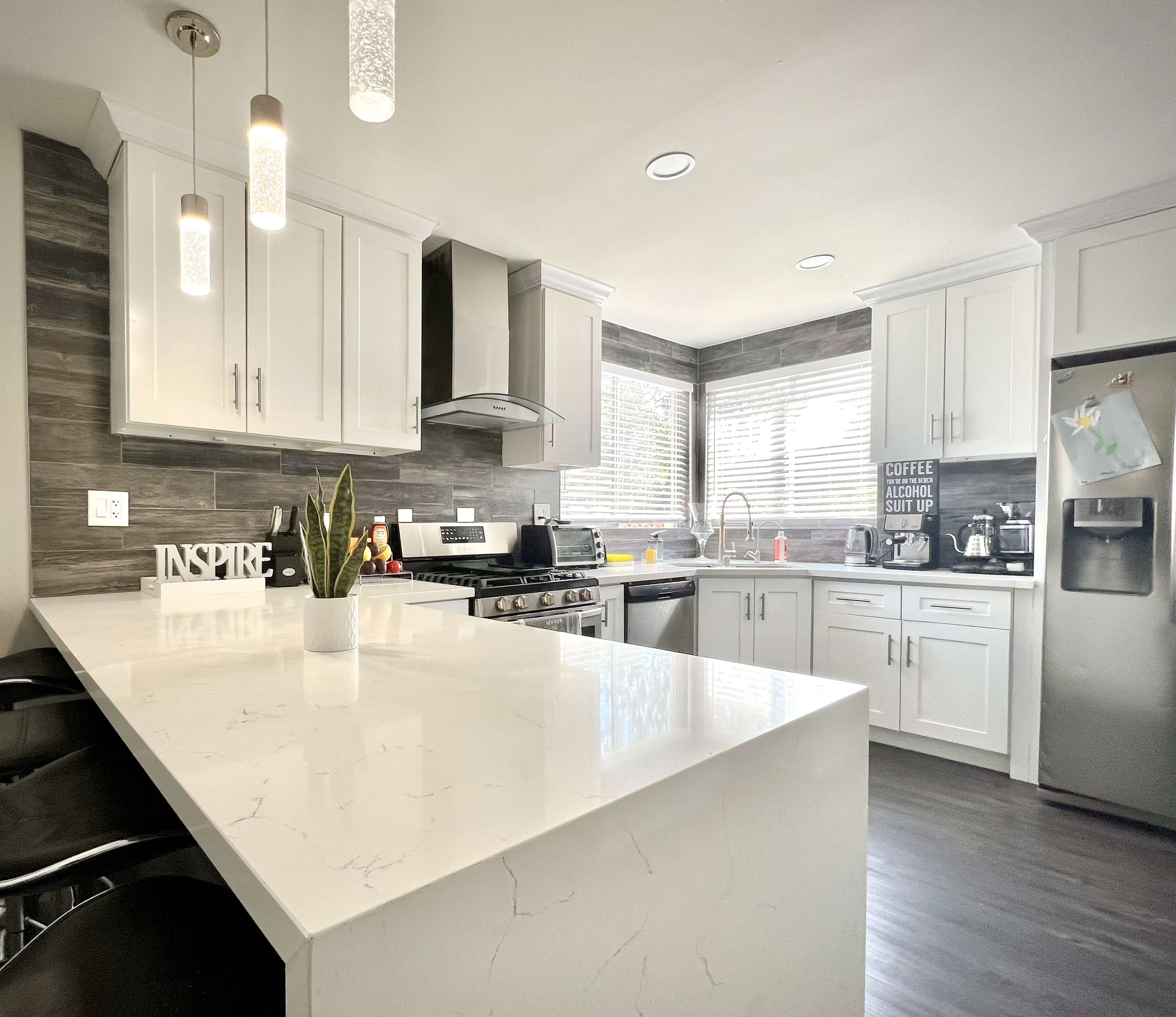 A modern kitchen features white cabinetry, a large island with a countertop, stainless steel appliances, and natural light streaming through the windows.