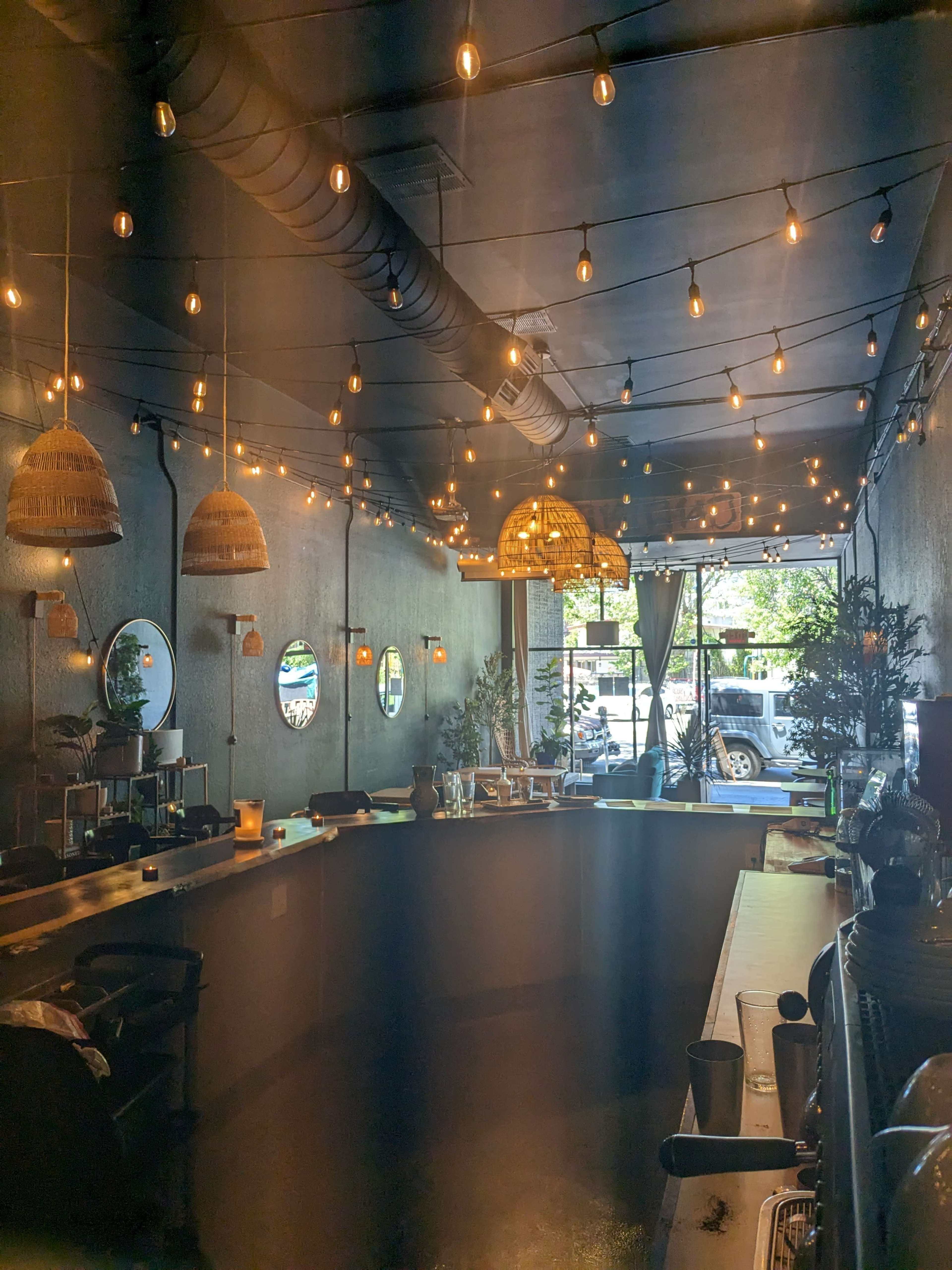 An interior of a café with round mirrors, hanging light fixtures, and plants, illuminated by string lights above a curved bar.