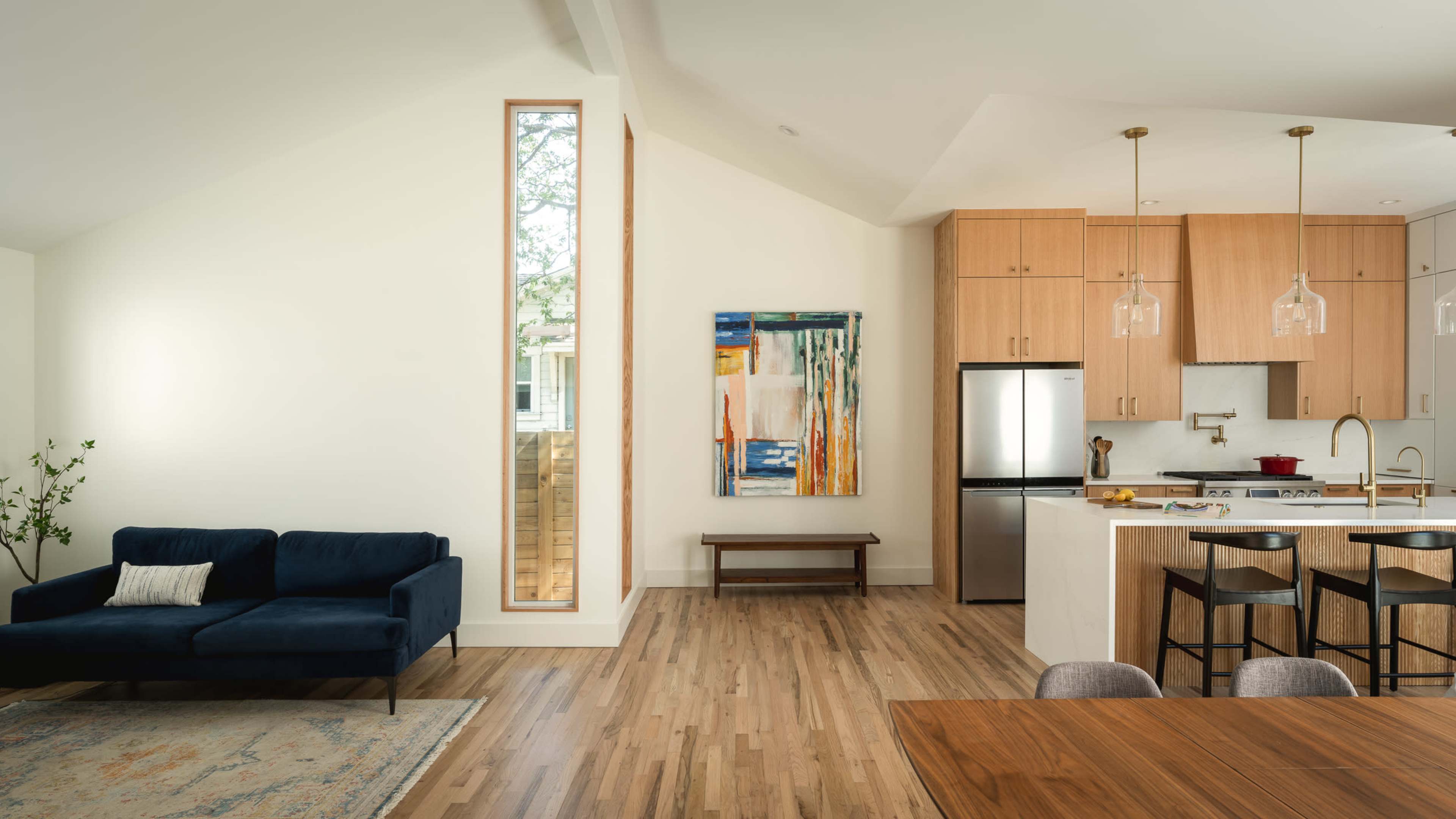 A modern living space features a dark blue sofa, a wooden dining table, and a kitchen with light-colored cabinets and stainless steel appliances, illuminated by natural light from a tall window.