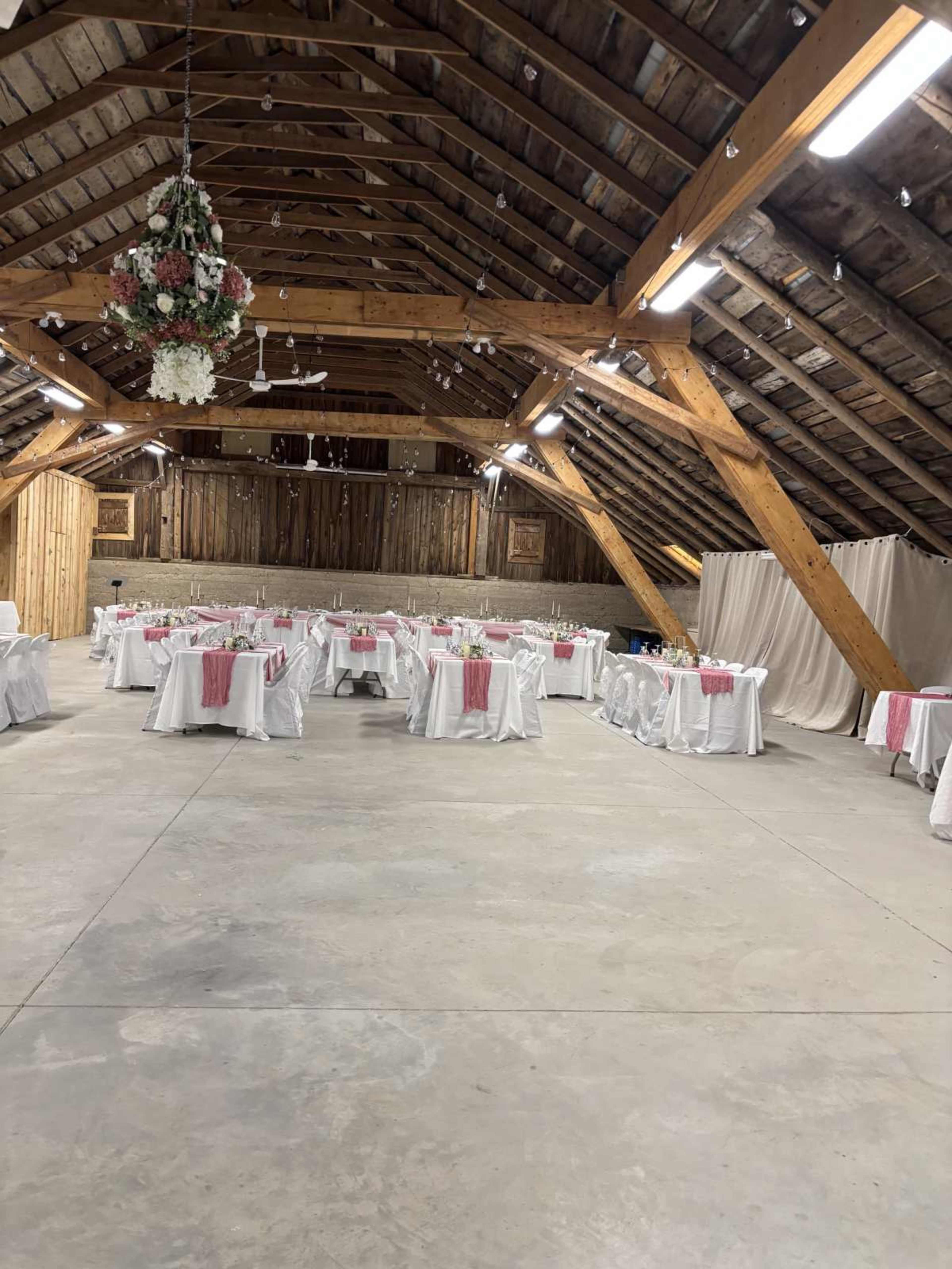 The image shows a rustic banquet hall with multiple tables dressed in white tablecloths and pink accents, arranged under a wooden beam ceiling.