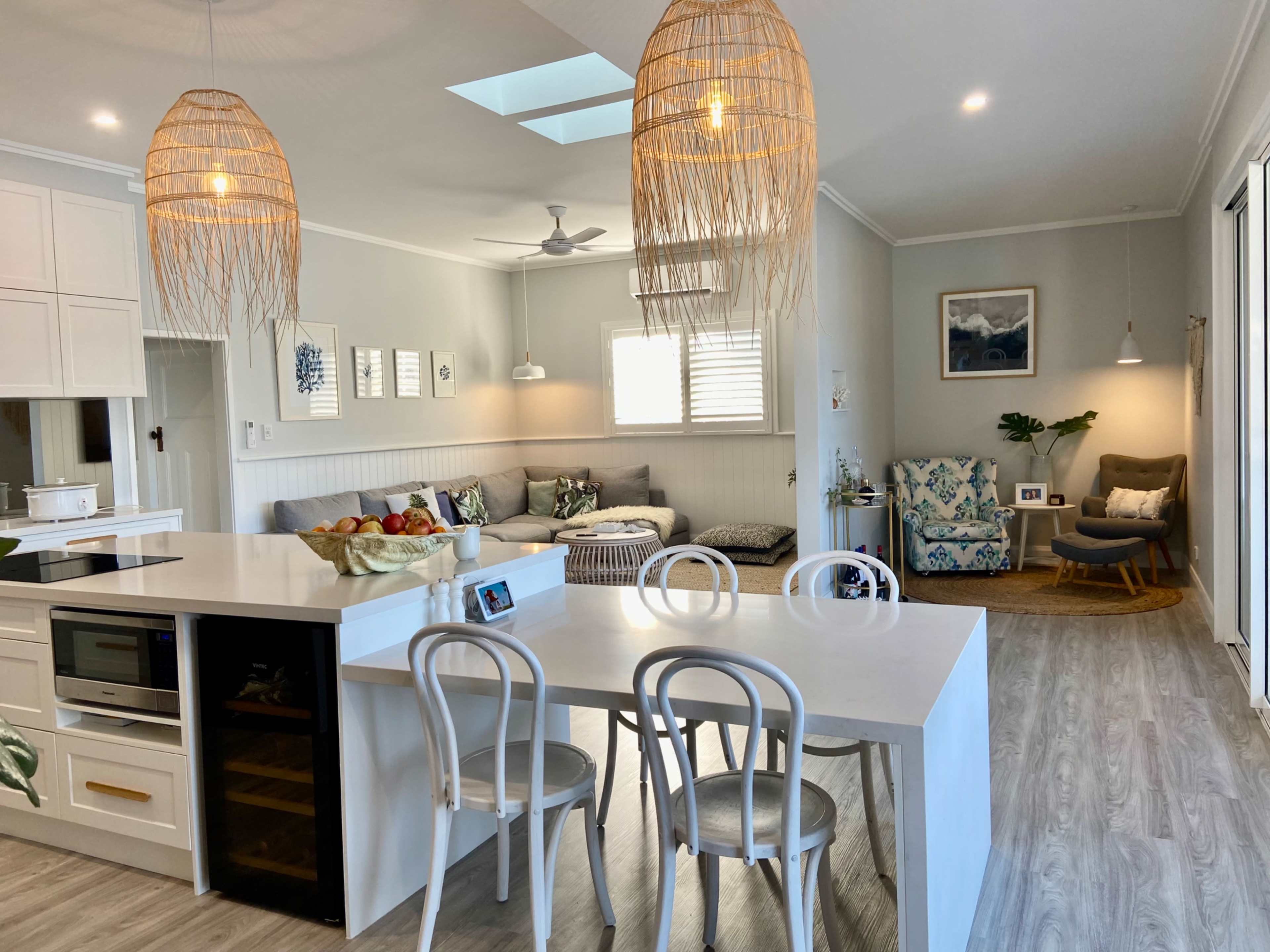 The image shows a bright, open-concept living space featuring a kitchen island with chairs, a comfortable seating area with decorative pillows, and natural light streaming in through a skylight.
