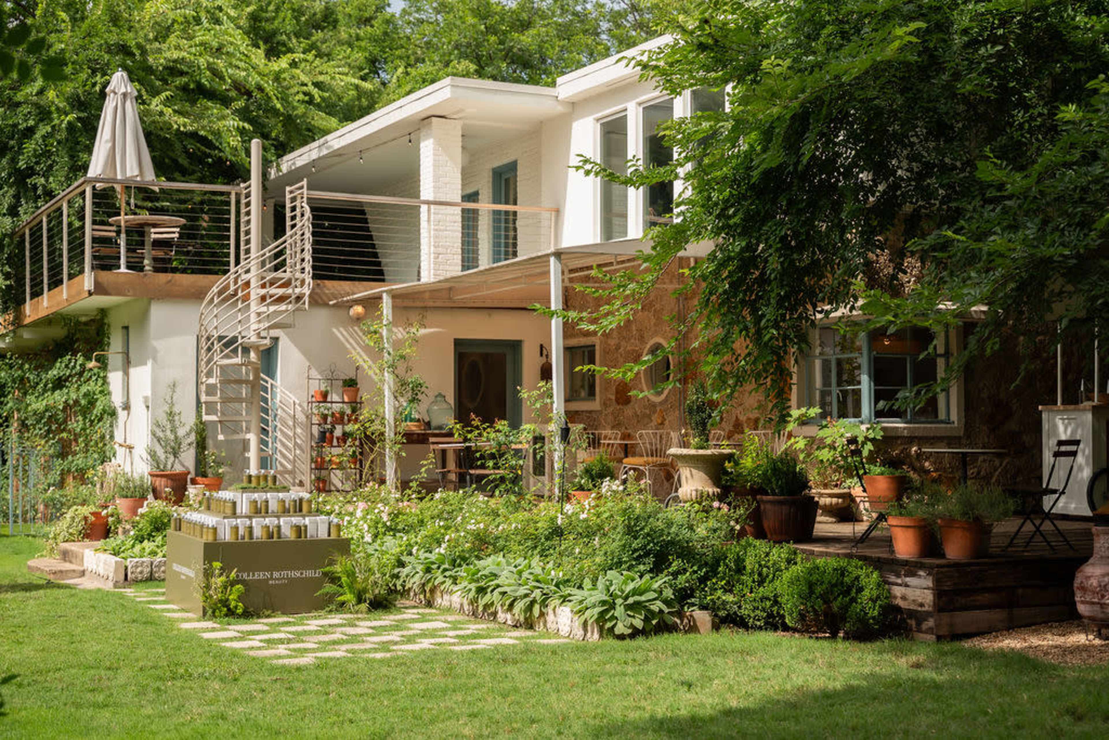 The image shows a modern home with a spiral staircase, surrounded by a garden filled with various plants and decorative pots.