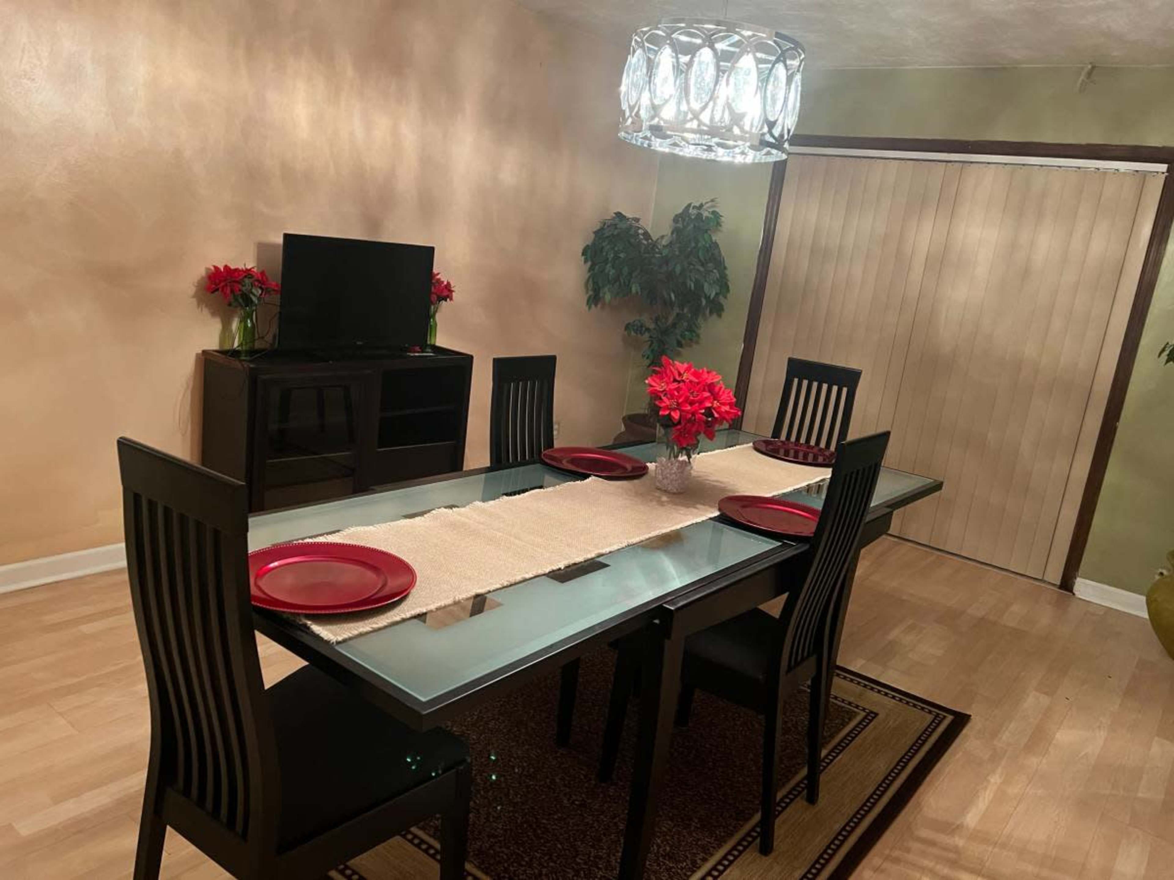A dining room features a glass table with six black chairs, red plates, a decorative centerpiece, and a television against the wall.