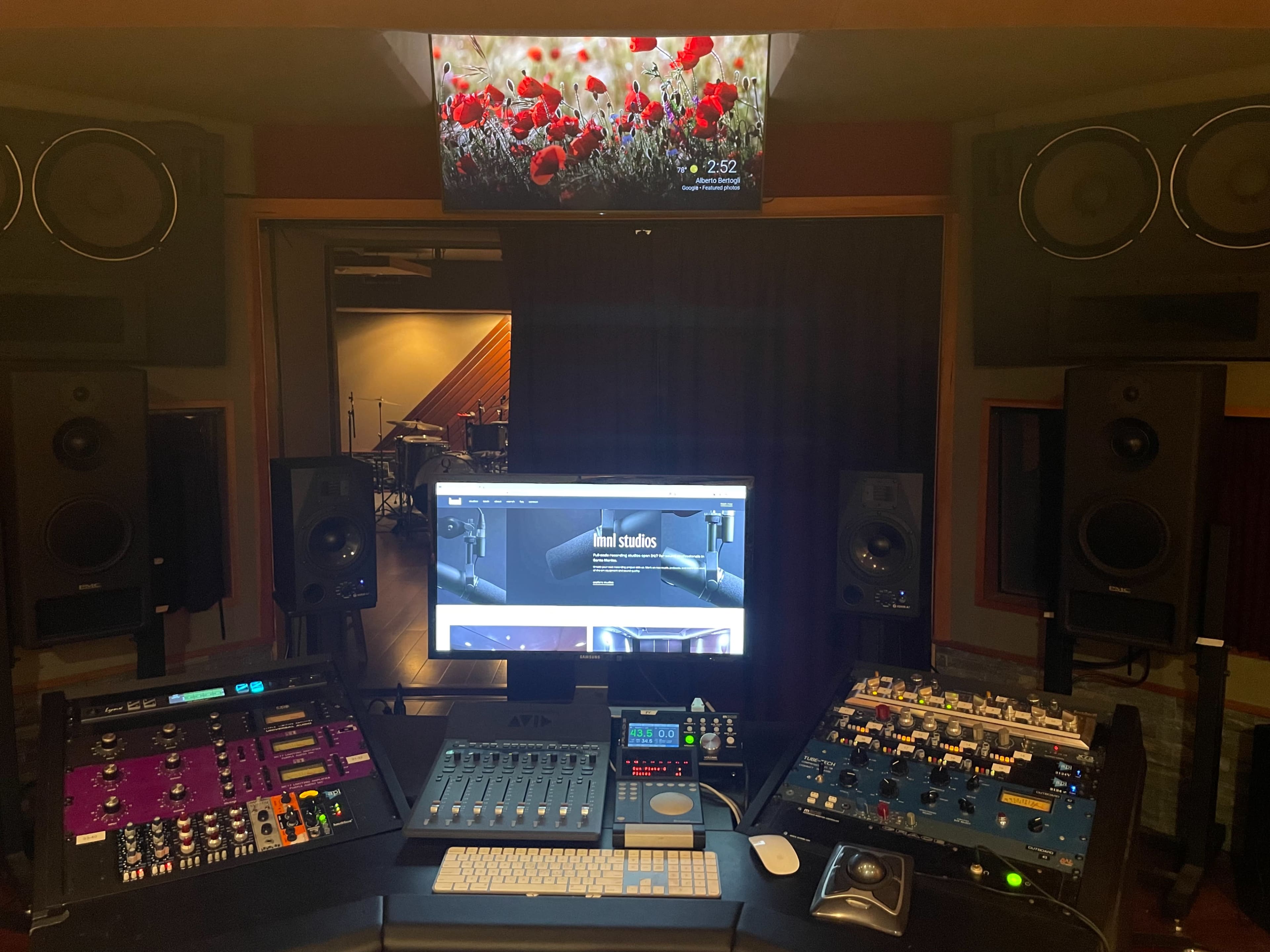 A music production studio featuring a central desk with audio equipment, a computer monitor displaying a website, and large speakers mounted on the walls.