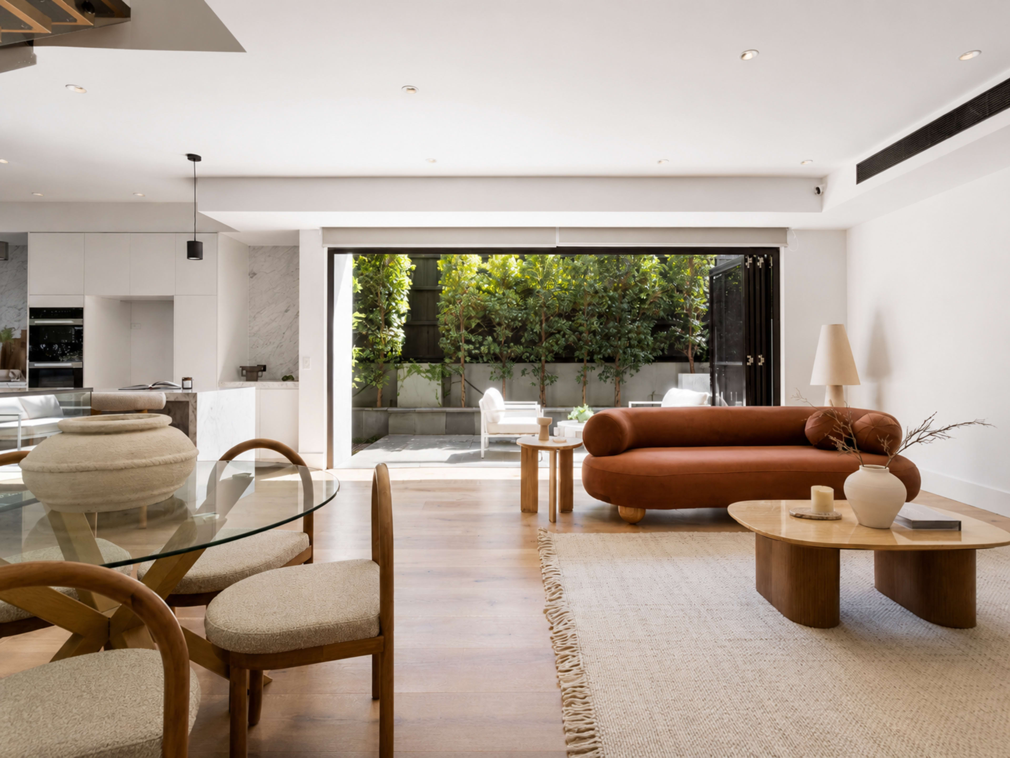 The image shows a modern living room with a round dining table, a brown curved sofa, and large glass doors opening to a green outdoor area.