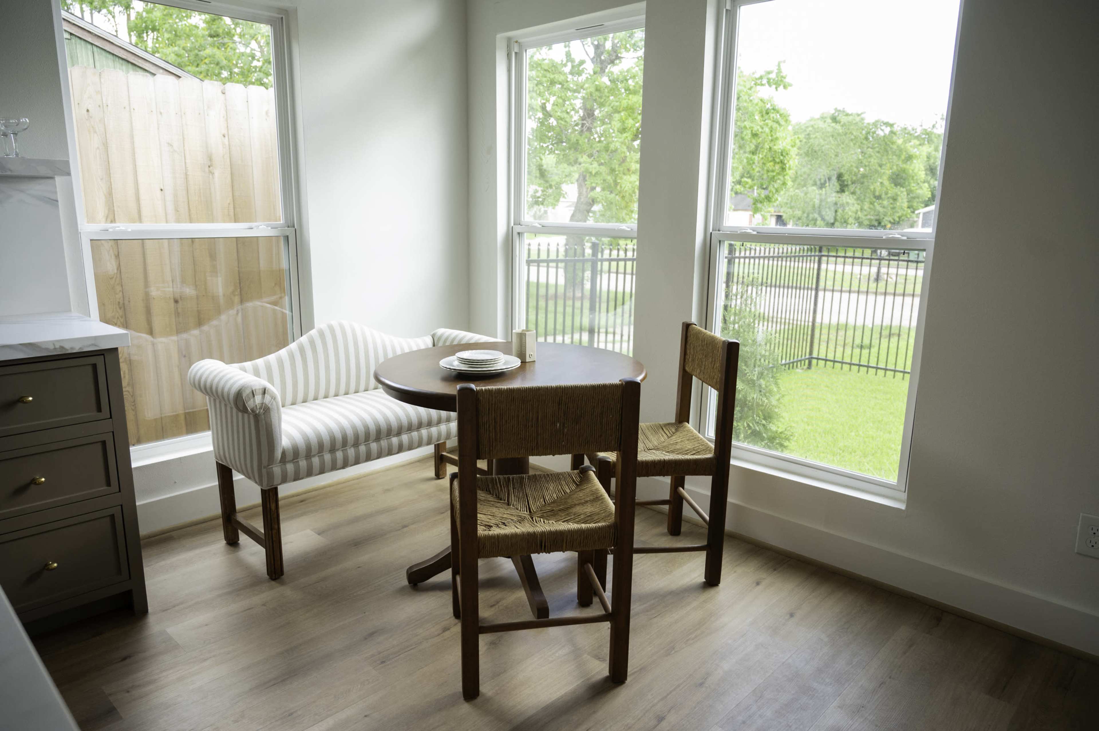 A small breakfast nook features a round wooden table with two chairs and a bench, surrounded by large windows with views of a green yard.