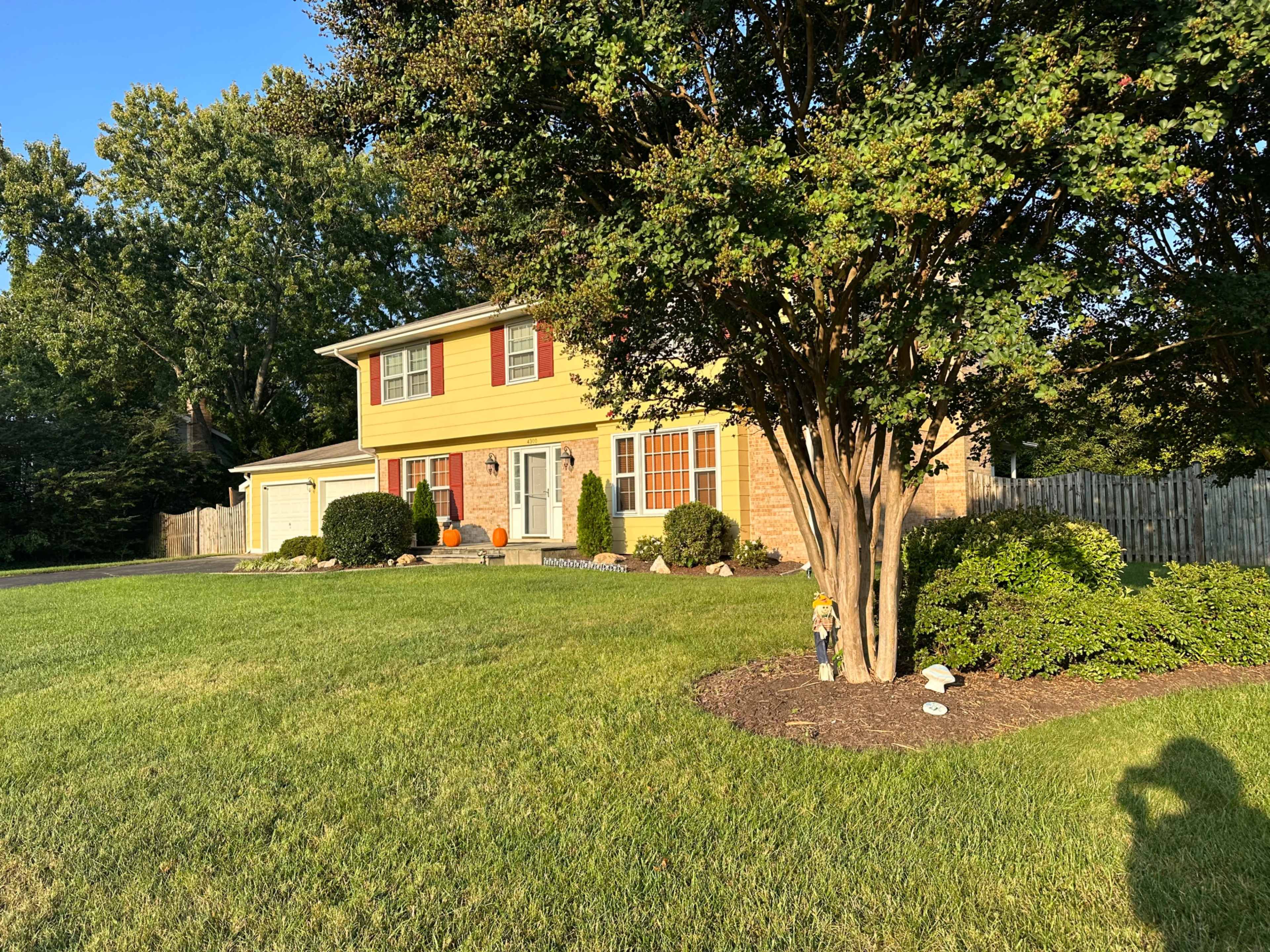 A two-story yellow house with red shutters sits on a well-maintained lawn, surrounded by trees and shrubs.