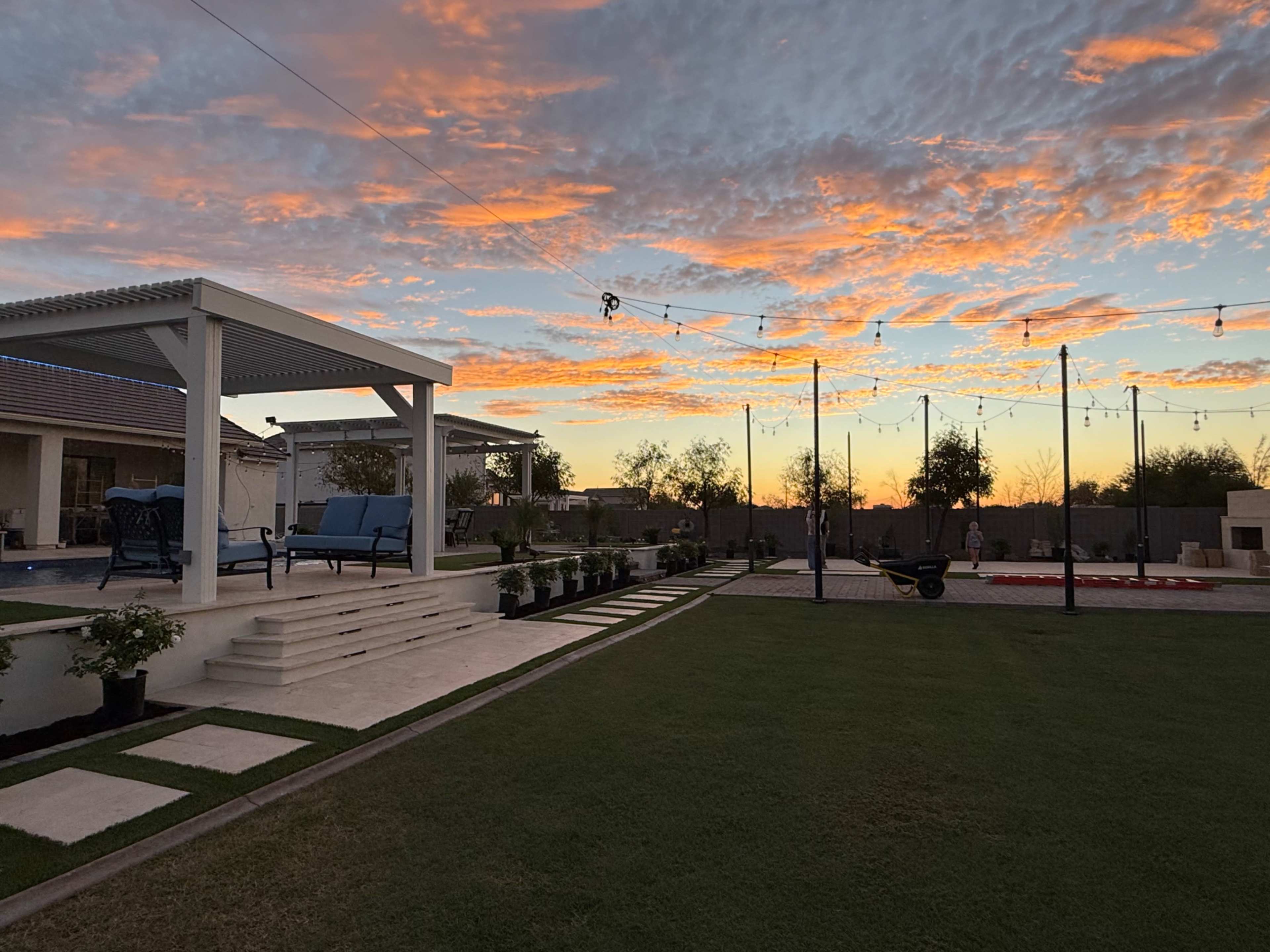 The image features a landscaped backyard with a seating area under a pergola, vibrant sunset clouds, and a grassy lawn.