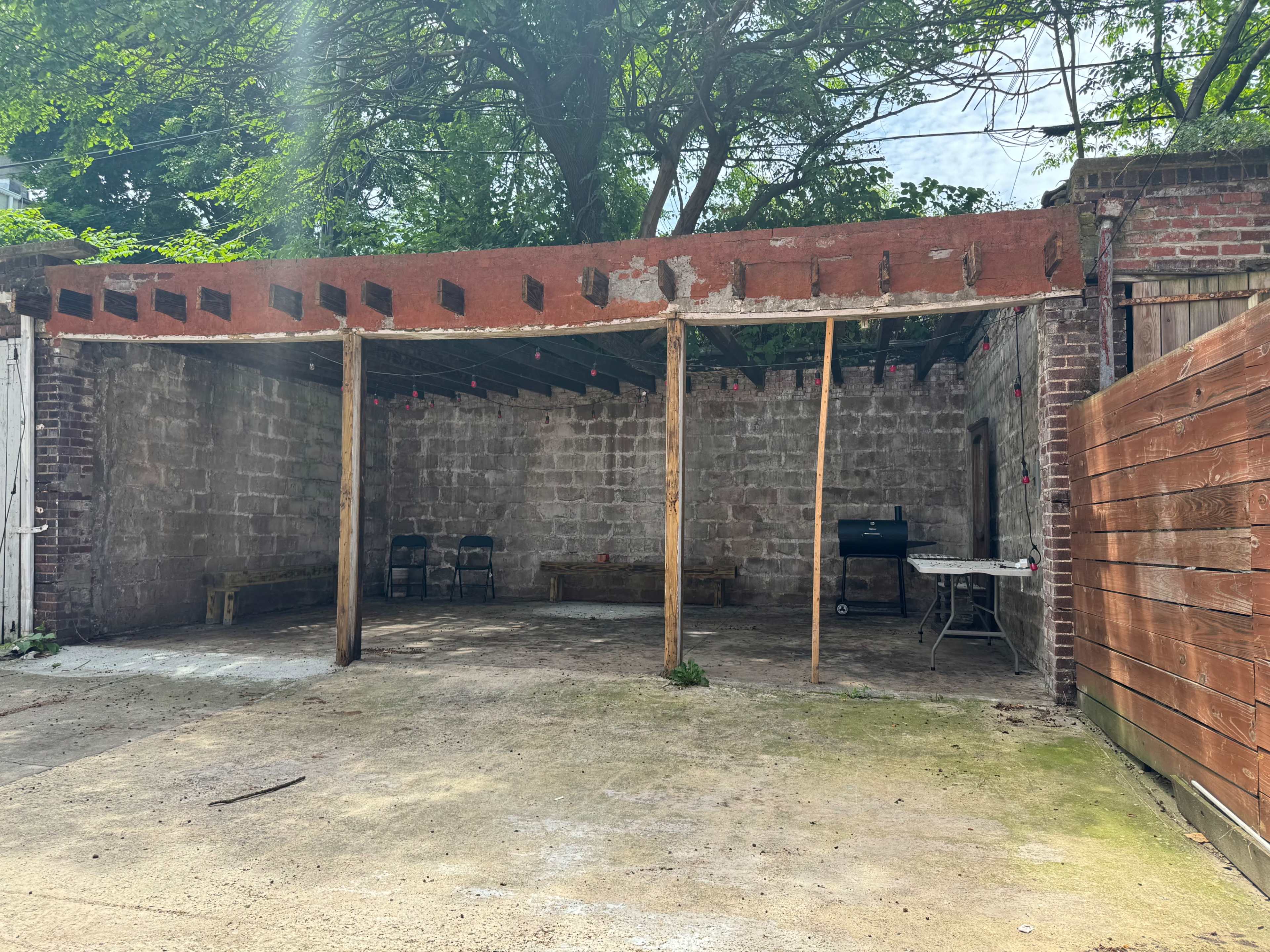 An unfinished, open outdoor area with a concrete floor, exposed brick walls, and wooden beams overhead.