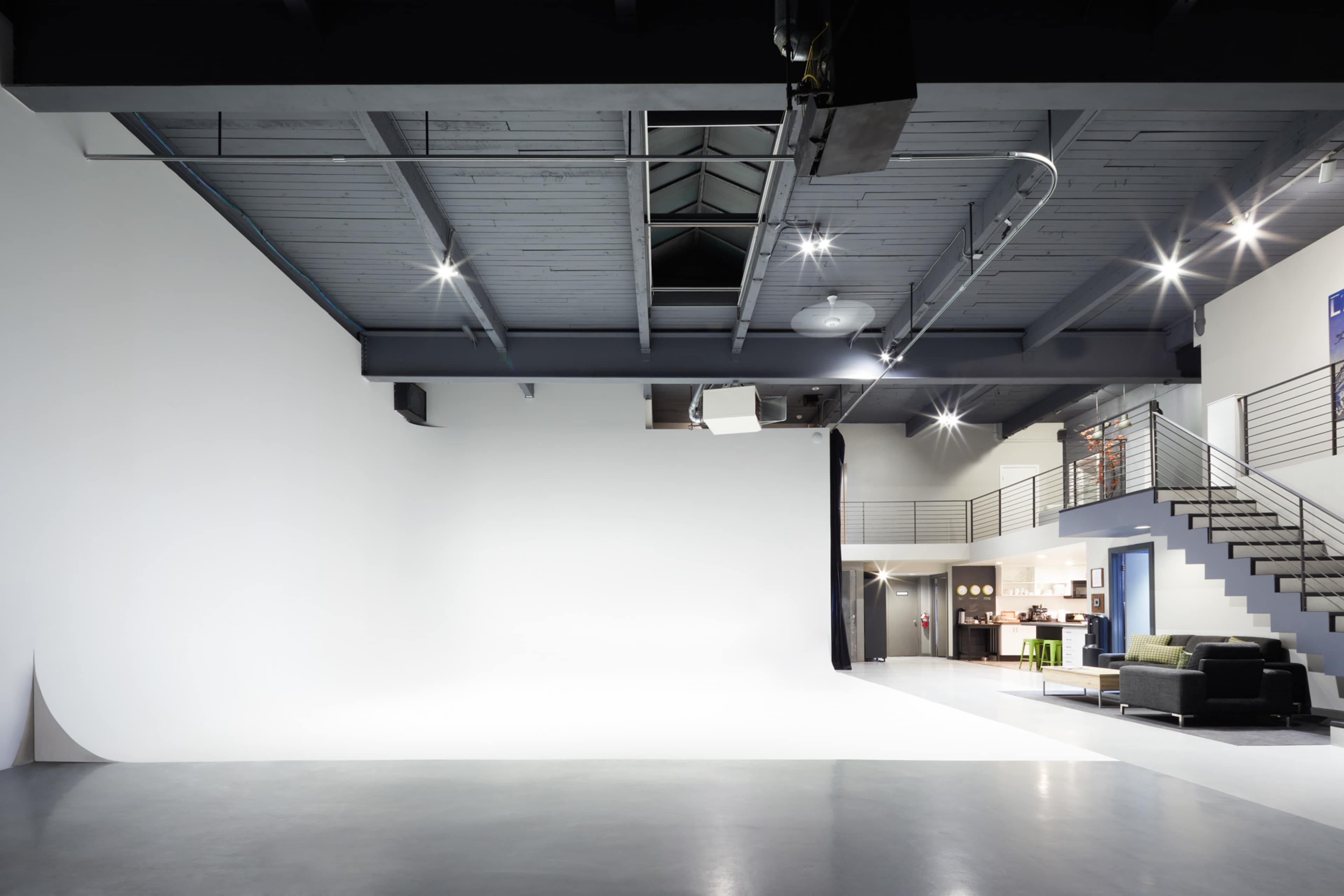 The image shows a spacious photography studio with a white backdrop, polished concrete floor, and a staircase leading to an upper level with a lounge area.