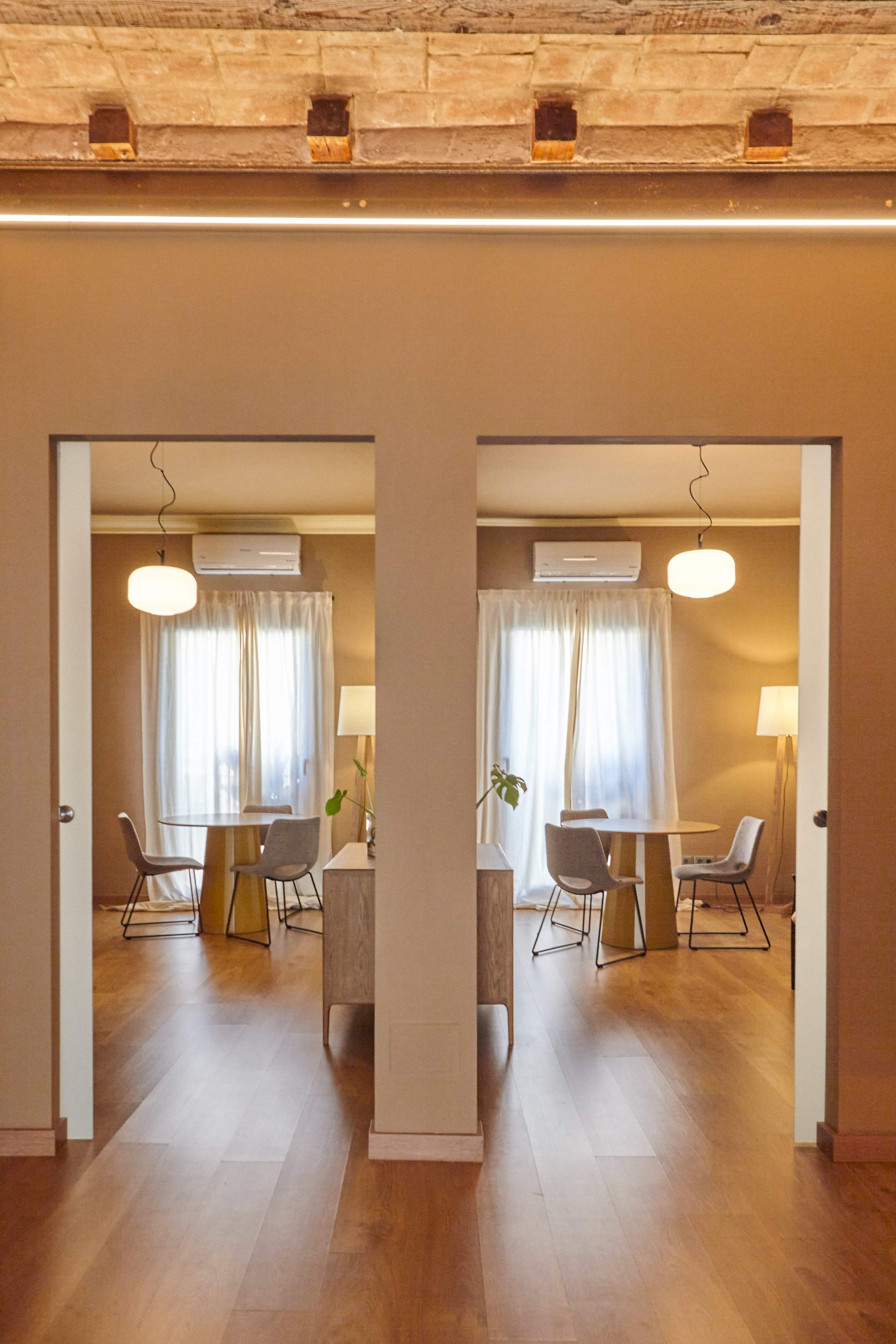 The image shows a corridor with two open doorways leading to two identical rooms, each furnished with a table, chairs, and lighting, and featuring large windows with sheer drapes.