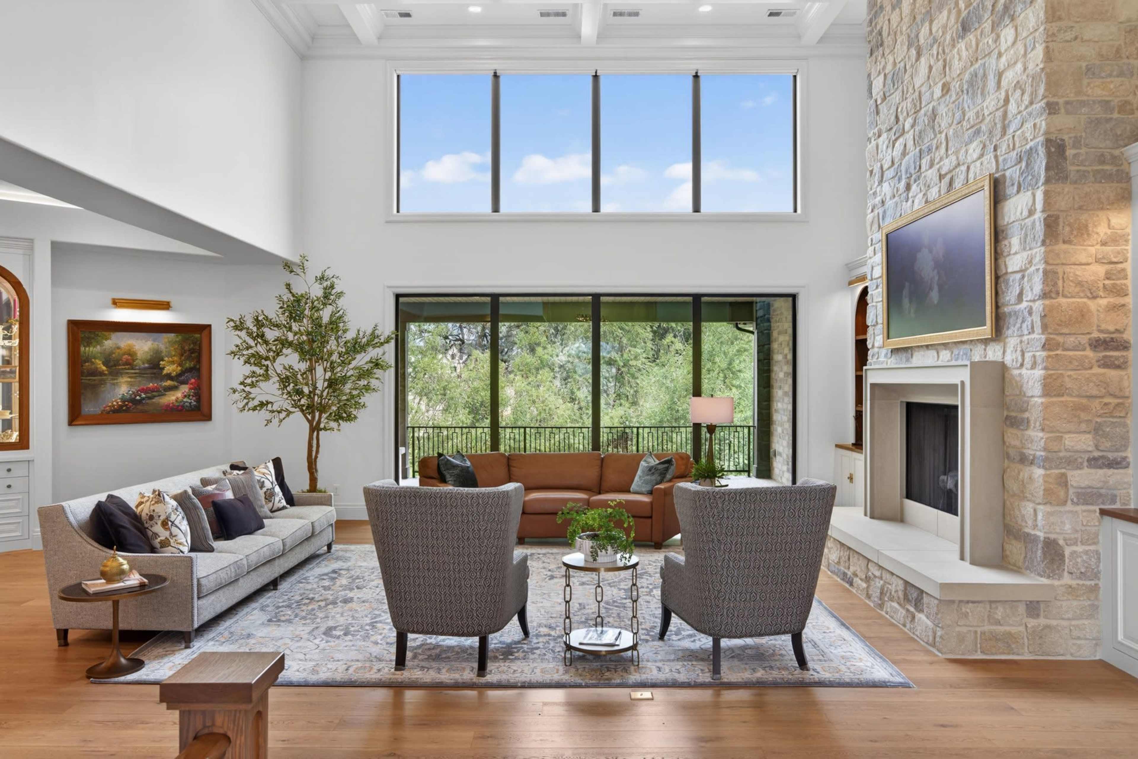 The image showcases a modern living room with a stone fireplace, large windows offering a view of greenery, and a combination of gray and brown furniture.