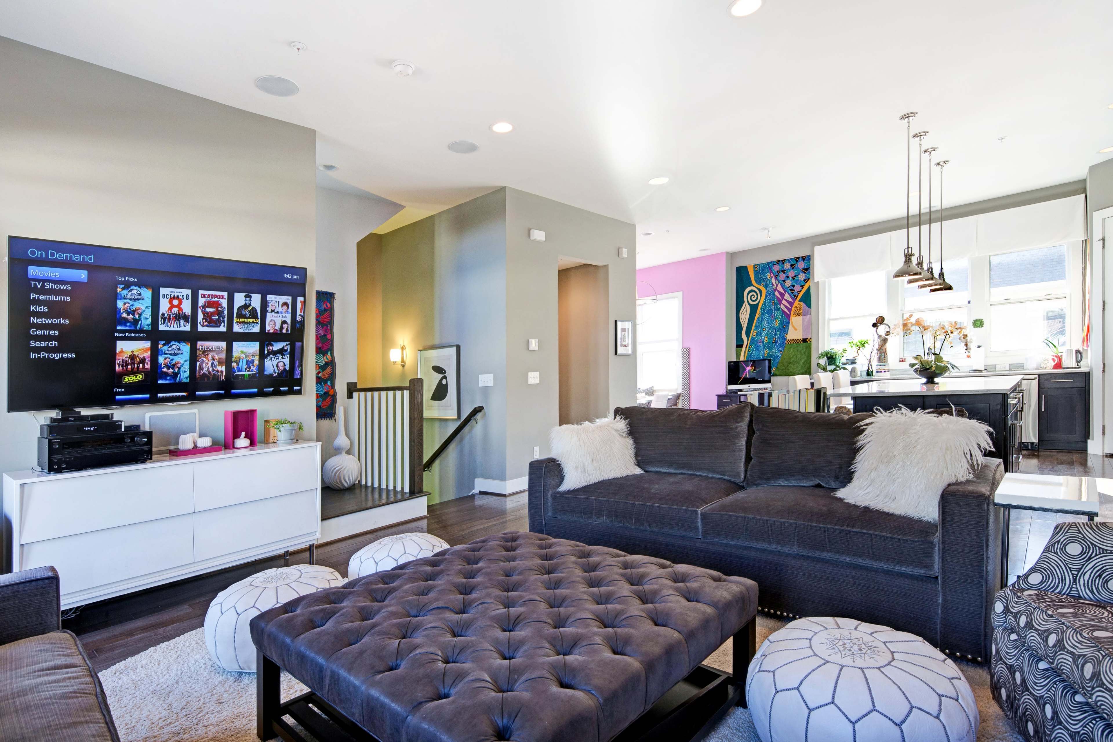 The image shows a modern living room featuring a large gray sectional sofa, a tufted coffee table, entertainment unit with a TV, and an open kitchen area in the background.