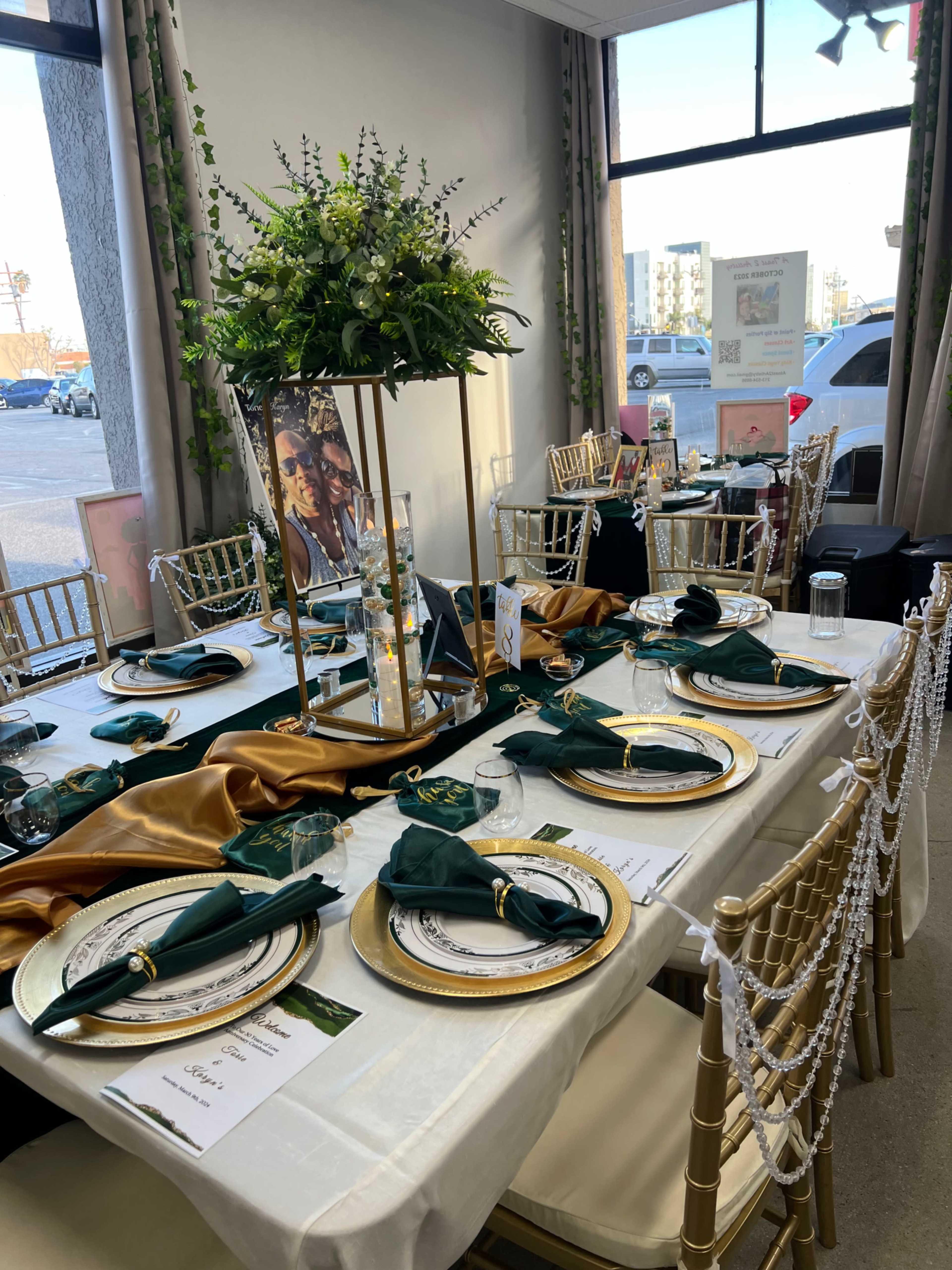 A elegantly arranged dining table features gold and green decorations, including plates, napkins, and a tall floral centerpiece, set against a window backdrop.