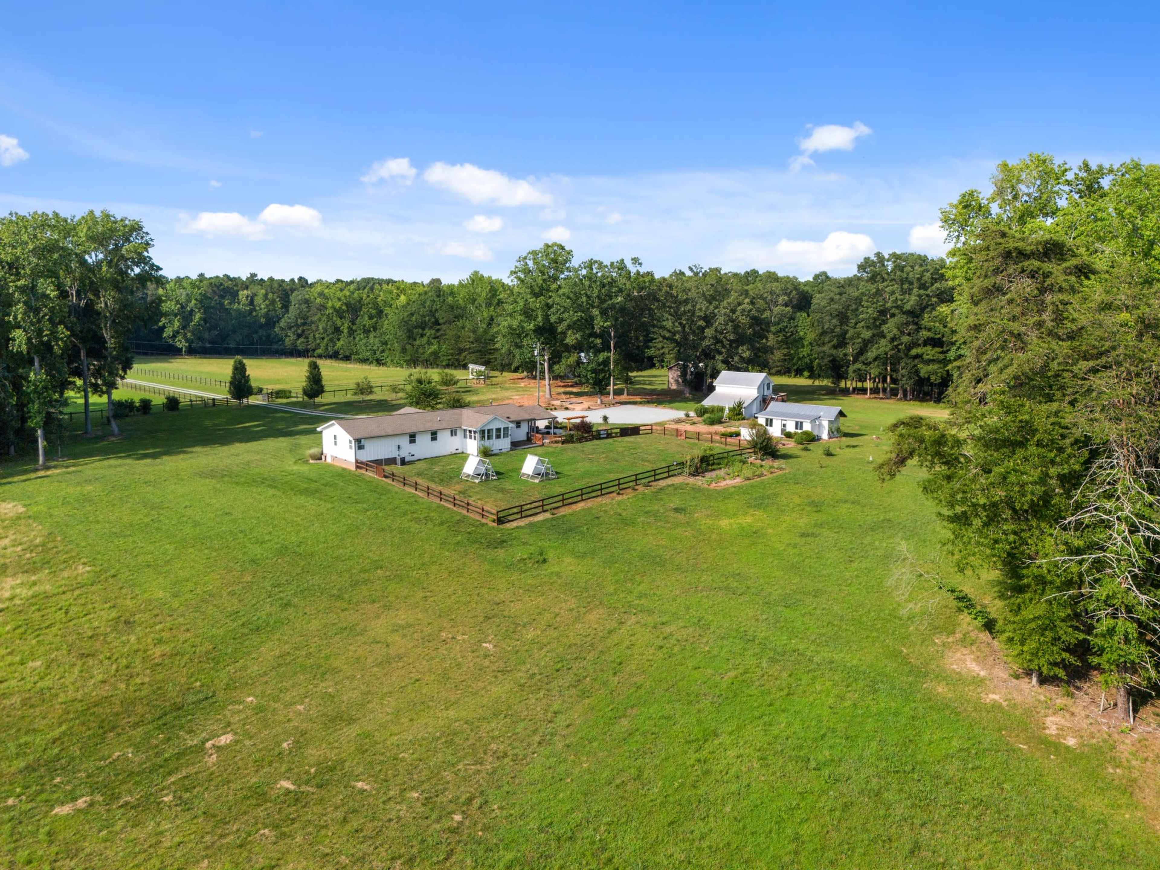 The image shows an aerial view of a spacious rural property featuring several buildings, fenced areas, and greenery, with trees in the background.