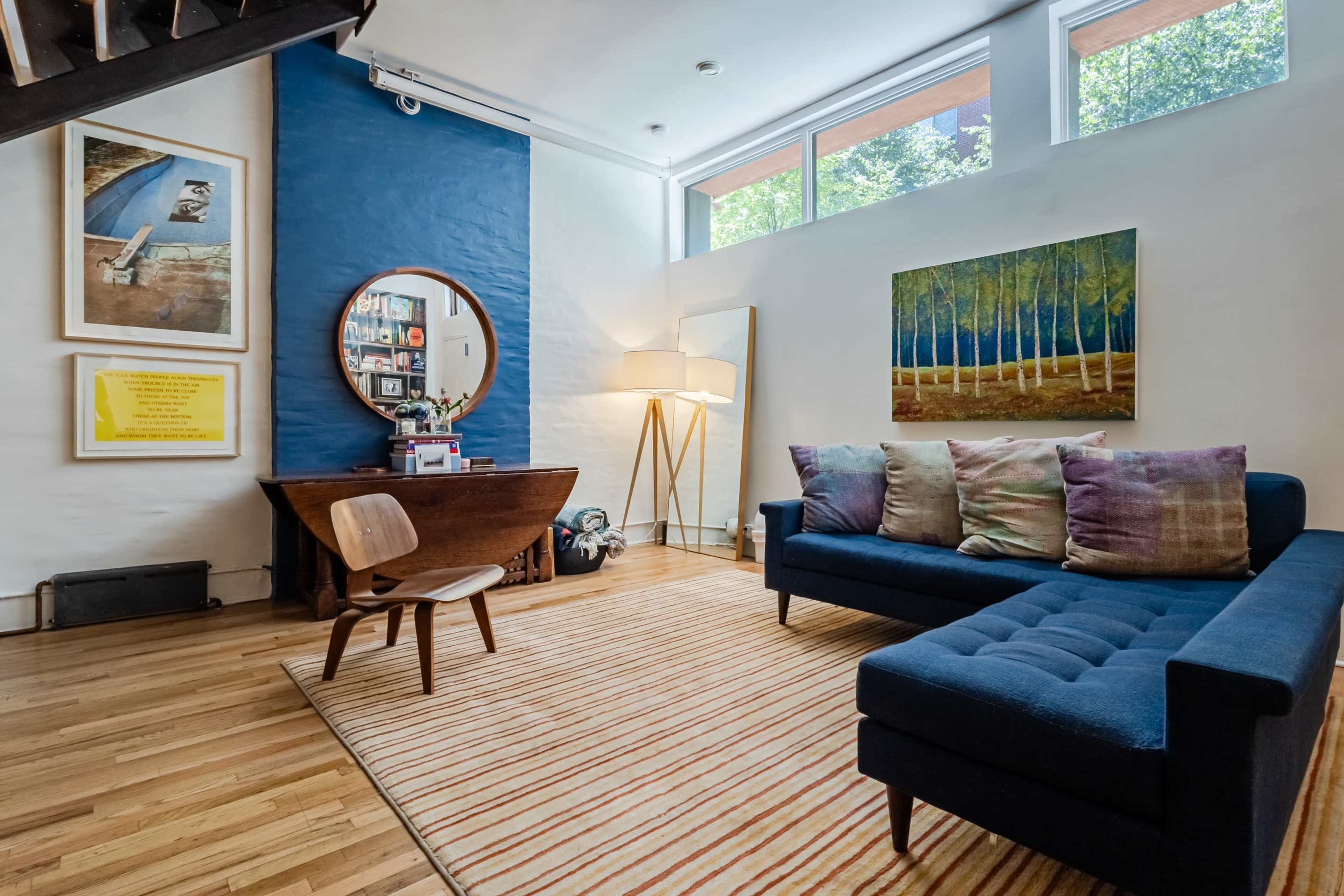 A cozy living room with a blue sofa, a wooden chair, a round mirror, and artwork on the walls, illuminated by natural light from large windows.