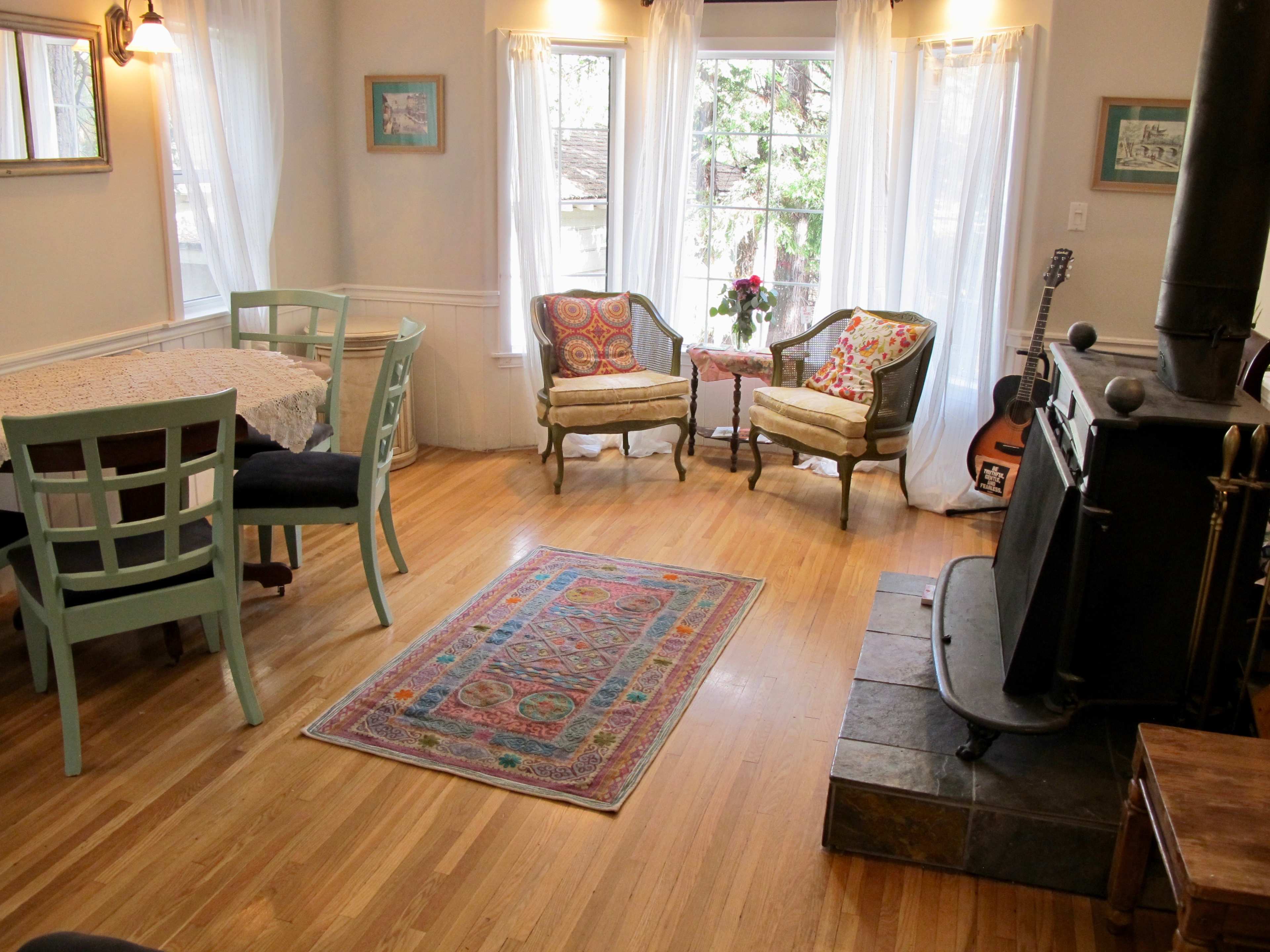 The image shows a cozy living space featuring a dining area with light blue chairs, a patterned rug, and two armchairs beside a window, along with a wood stove and a guitar.