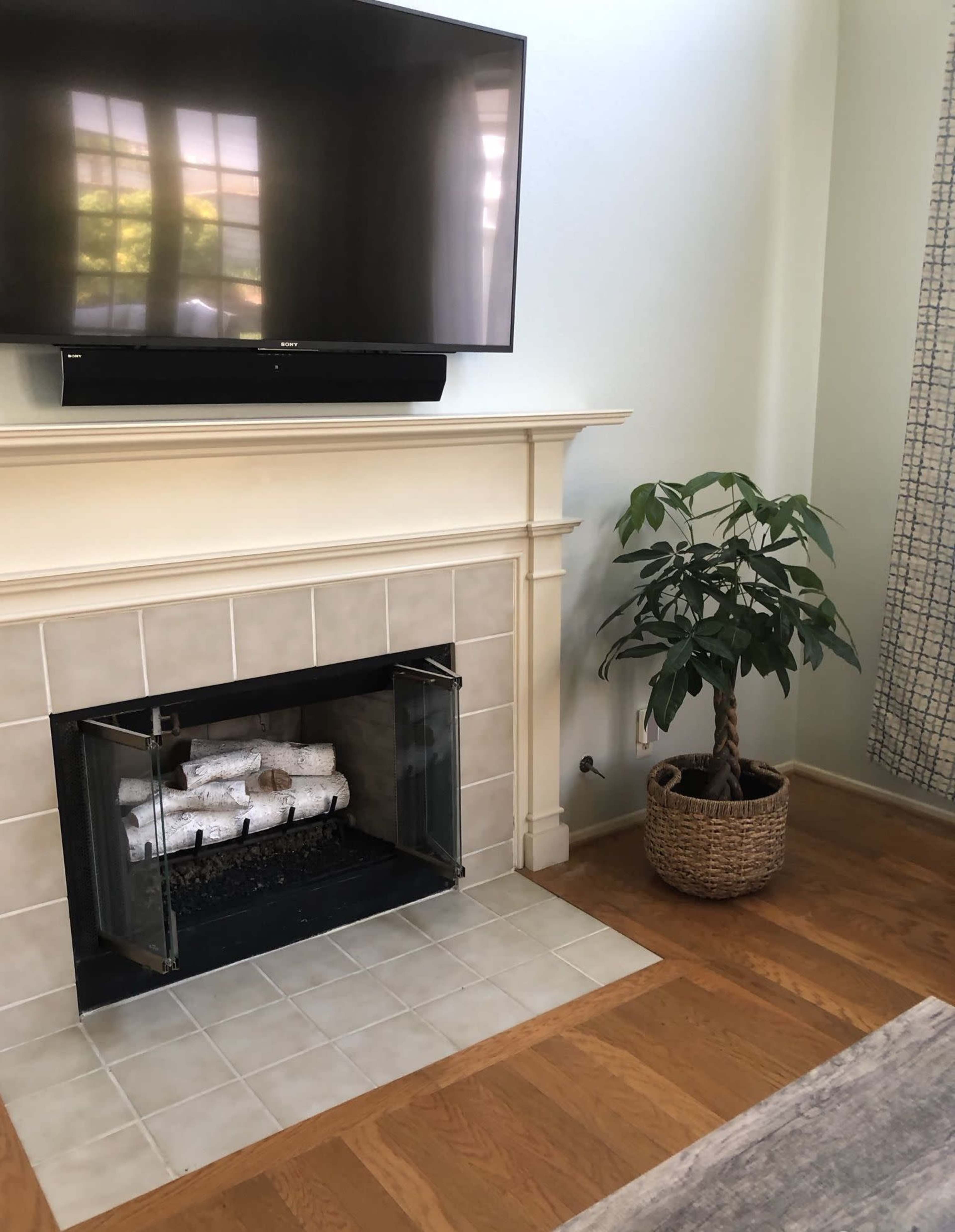 A living room corner with a wall-mounted television above a tiled fireplace and a potted plant in a woven basket nearby.