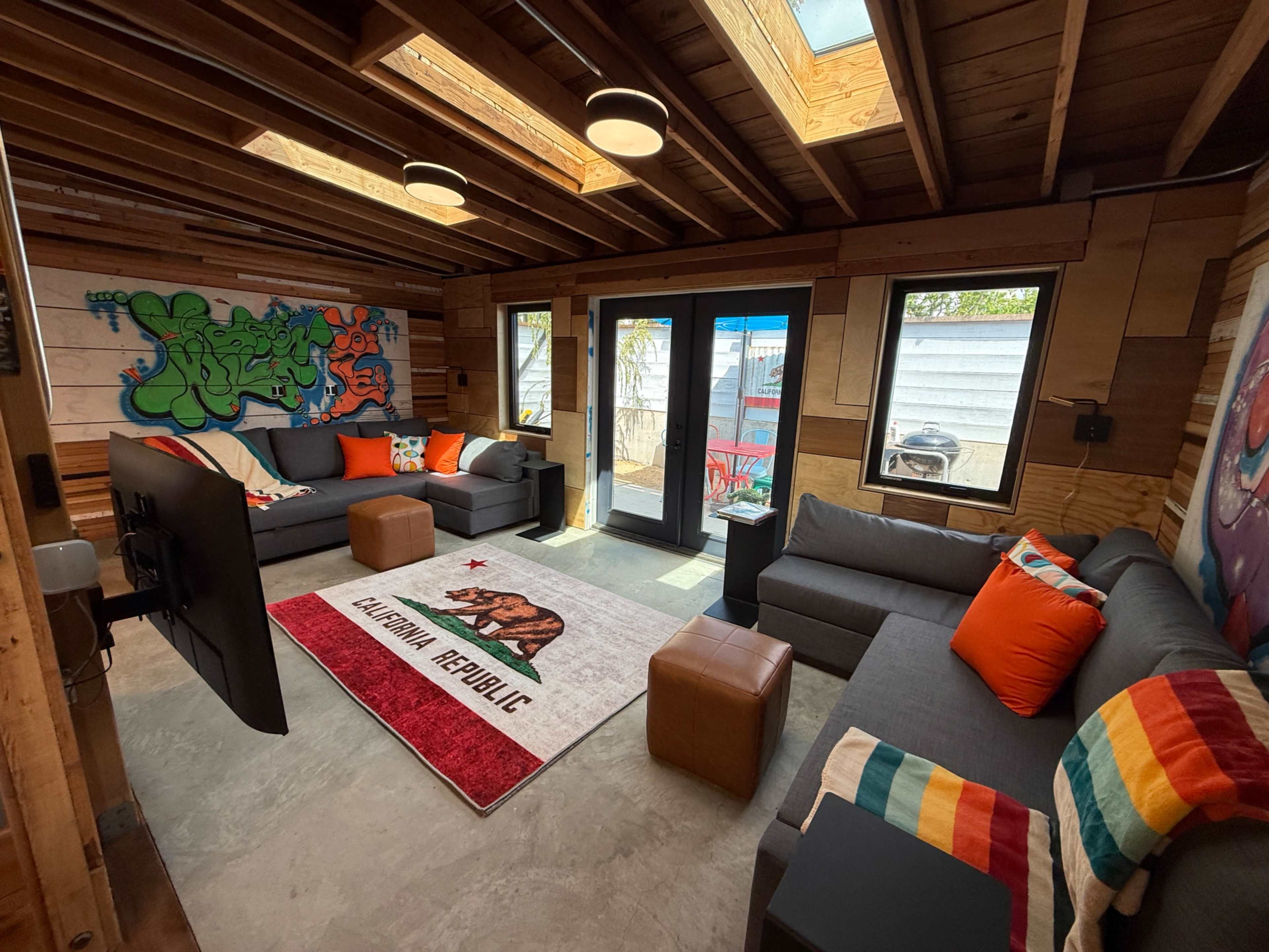 The image shows a modern, wooden-finished living room with gray sofas, bright cushions, a California bear rug, and large windows allowing natural light.