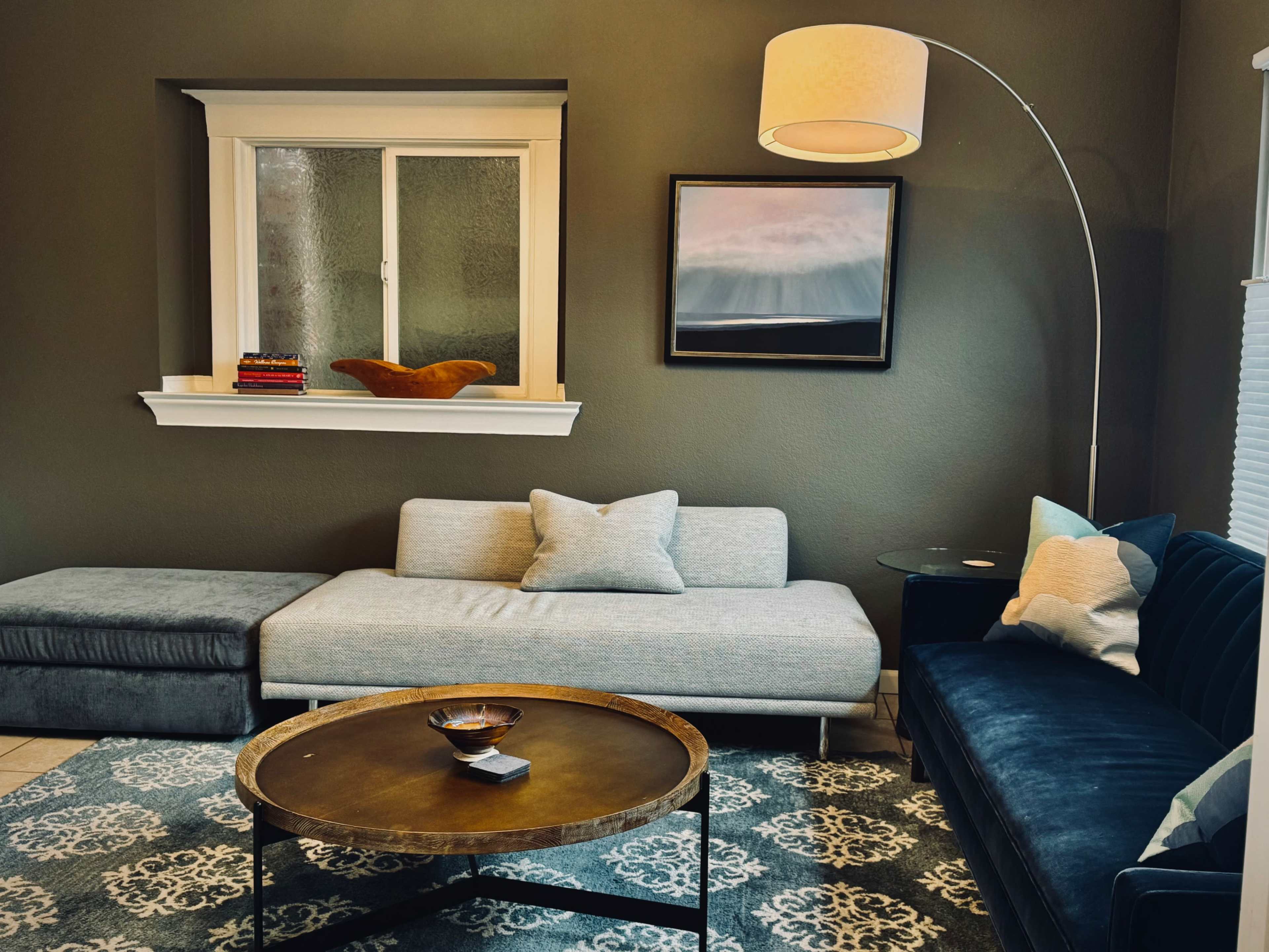 A modern living room features a gray sectional sofa, a round metal coffee table, and a floor lamp beside a dark blue couch, with a window and artwork on the walls.