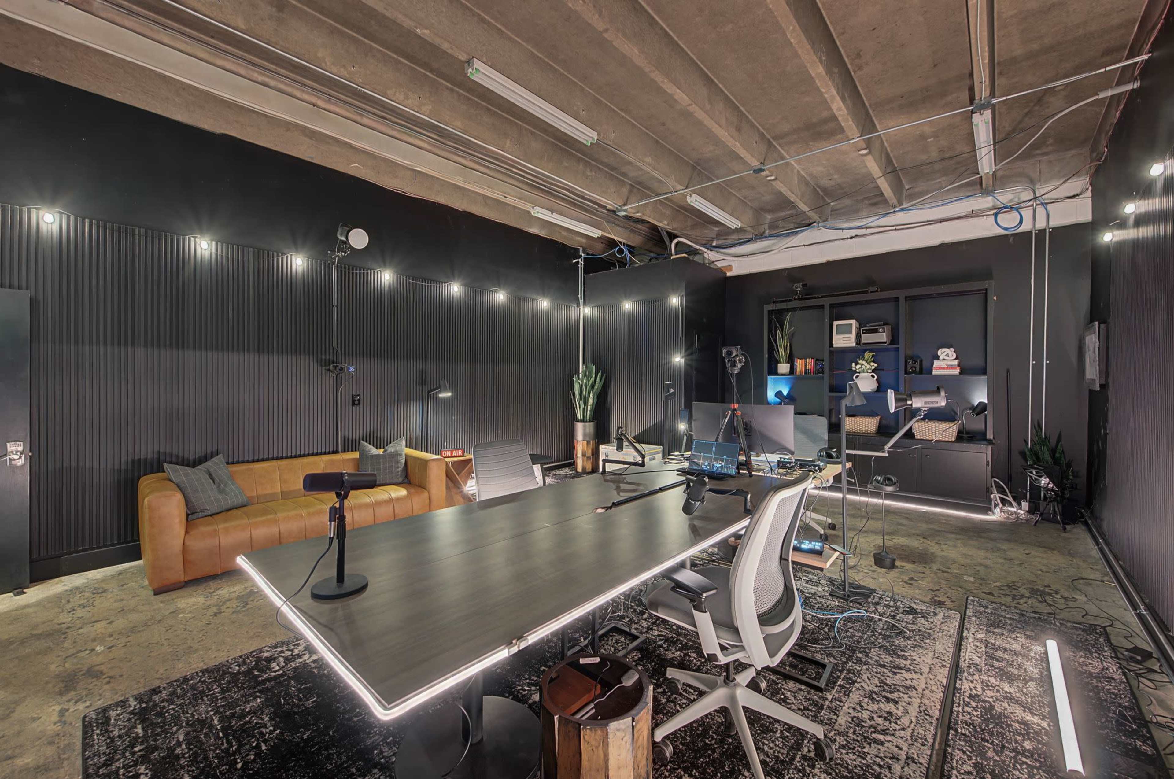The image shows a modern office space with a sleek desk, ergonomic chairs, a brown leather couch, and ambient lighting against dark walls.