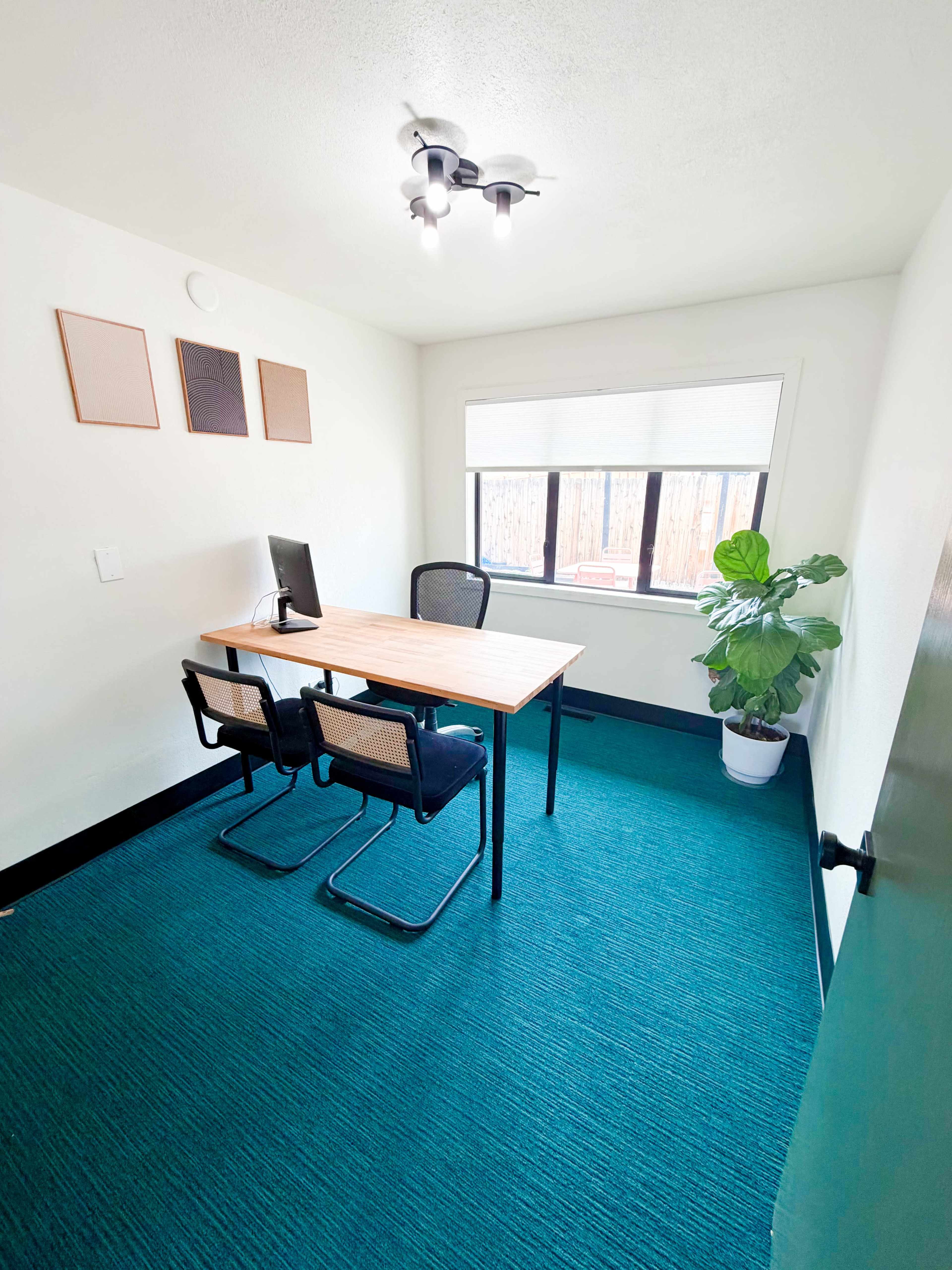 The image shows a small, minimalistic office space with a wooden desk, two chairs, a computer monitor, and a potted plant, all set against a teal carpet and white walls.